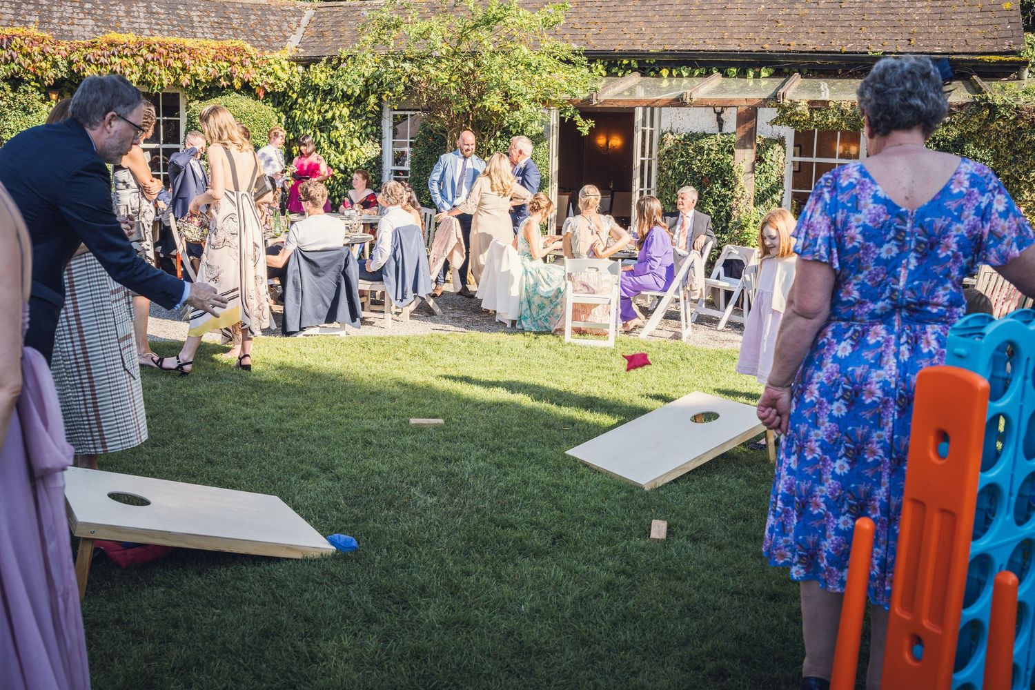 Wedding guests enjoy a lawn cornhole game while others watch from chairs at an outdoor reception.