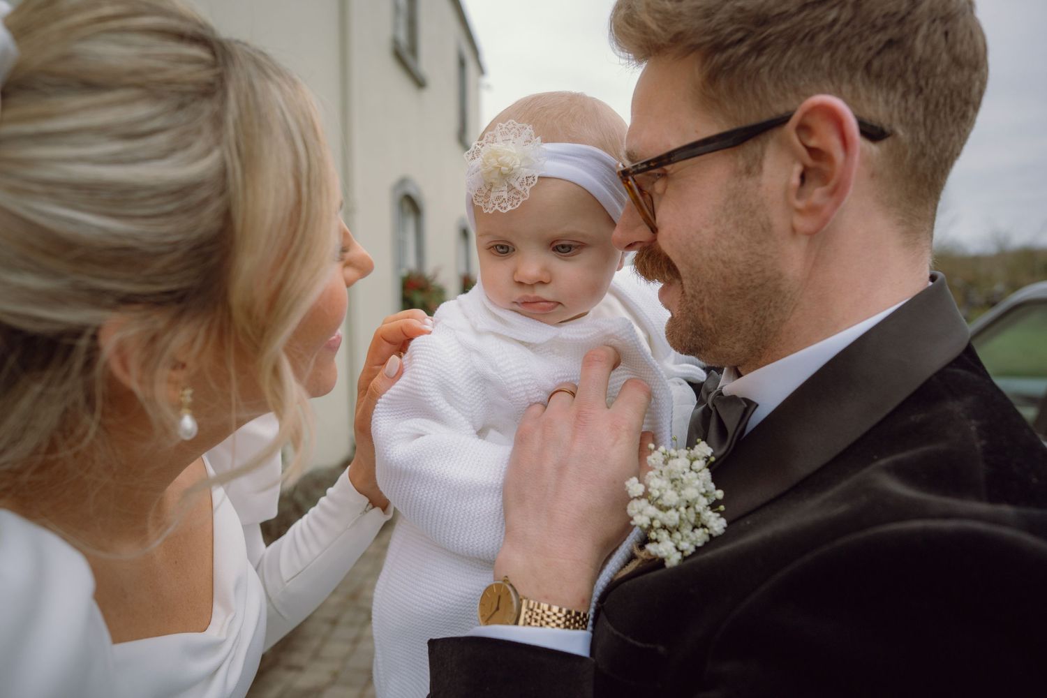 Newlywed couple lovingly tend to their baby dressed in white at outdoor wedding venue.