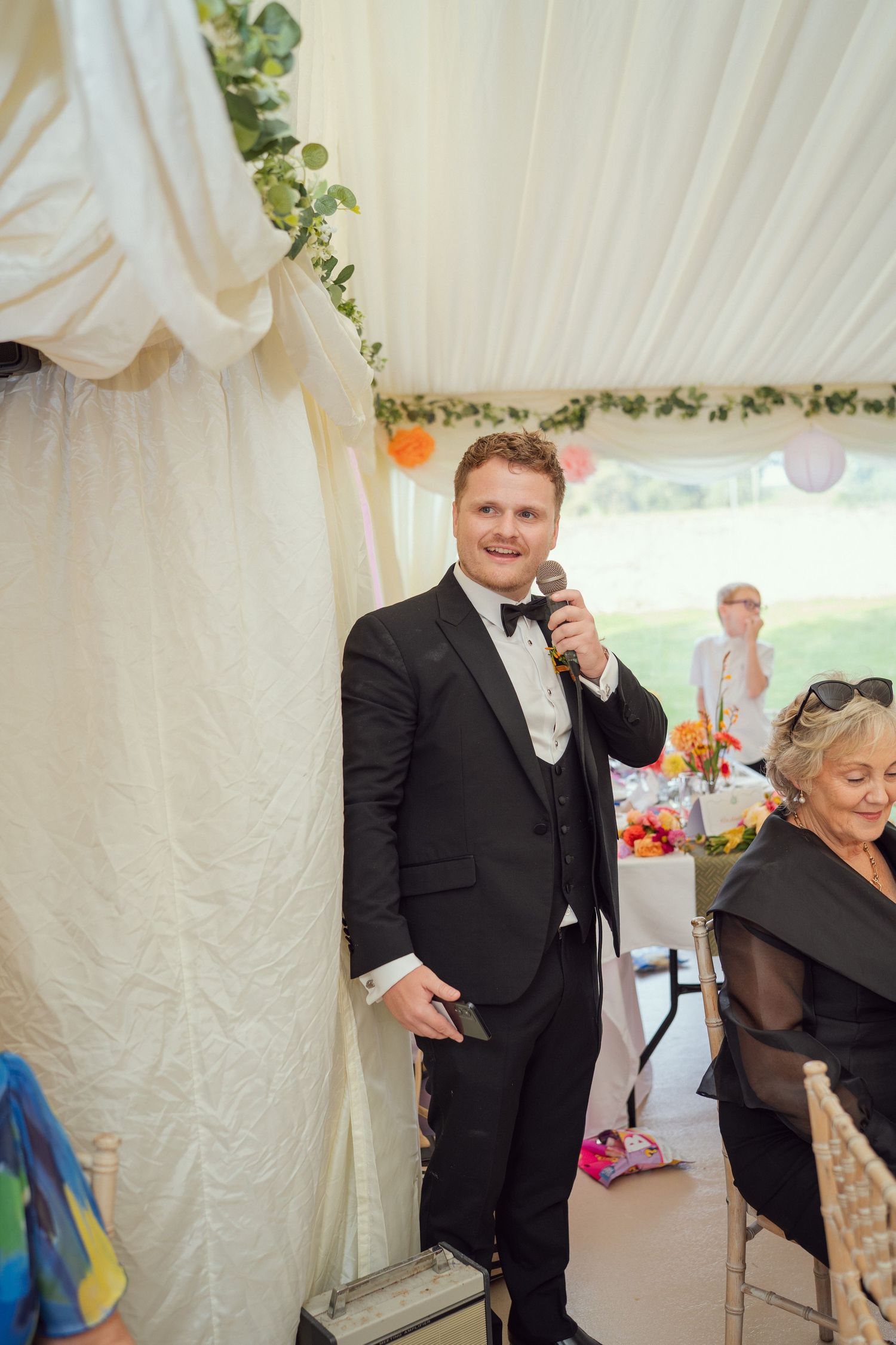 Brother of bride in black tuxedo giving a speech with a microphone at wedding reception at outdoor garden wedding reception in Co Meath.