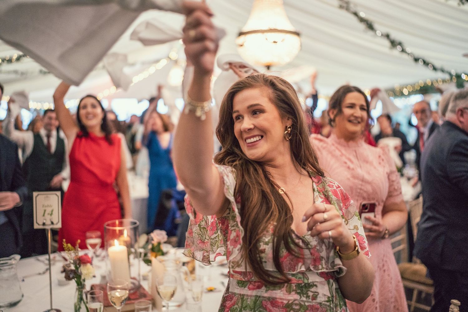 Joyful wedding guest in floral dress raising a glass and celebrating on the dance floor at a marquee reception.