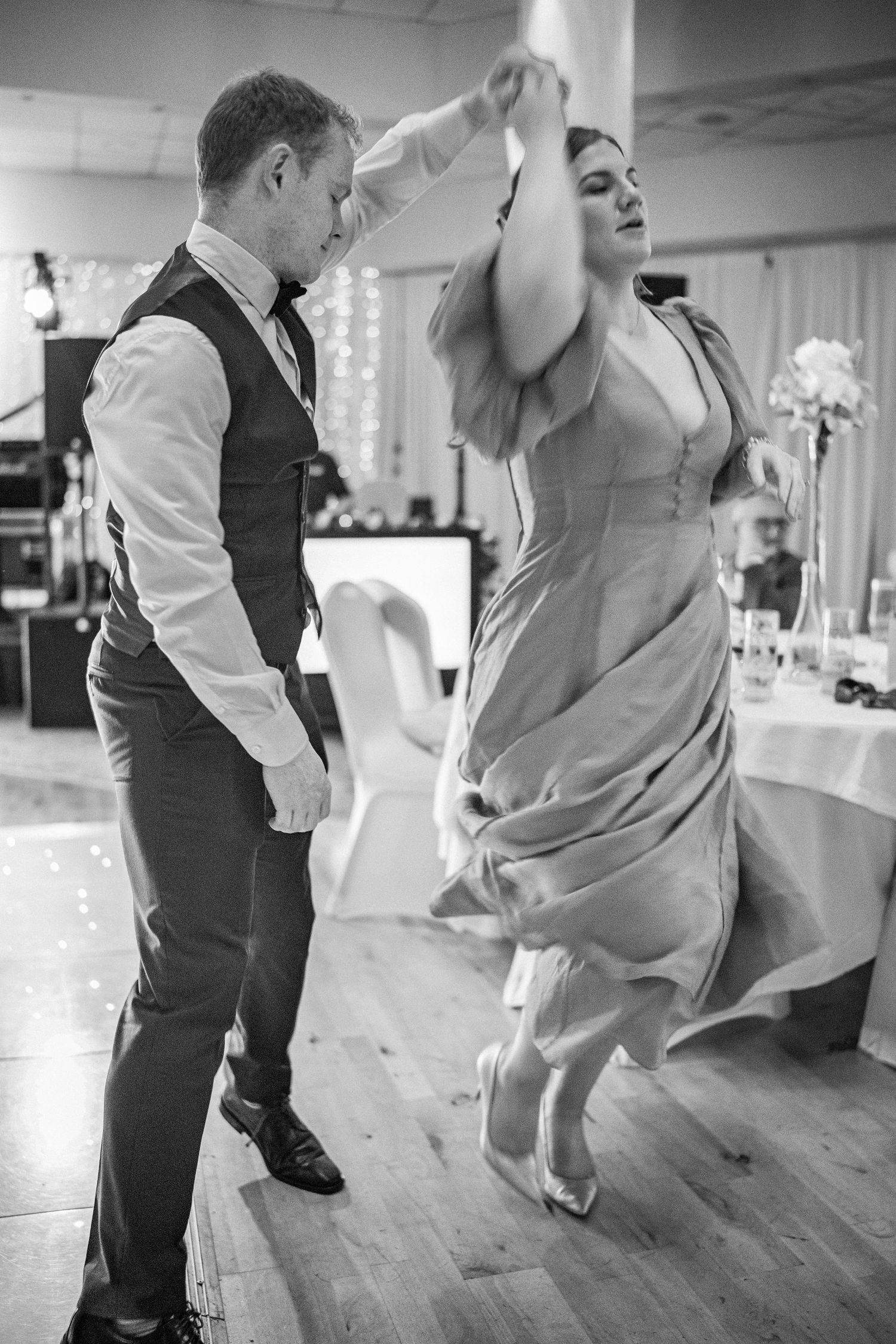 Couple dancing energetically at wedding reception, man spinning woman in elegant flowing gown.