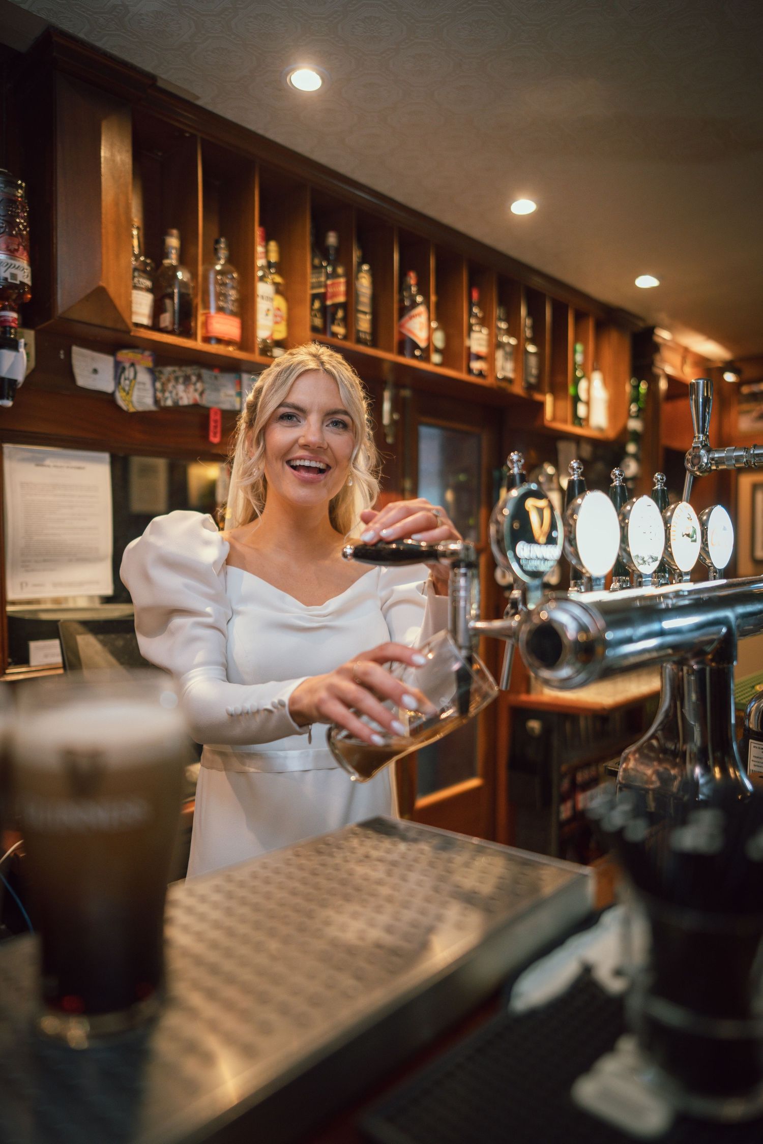 Radiant blonde bride in white dress pours Guinness at a traditional wooden pub bar, smiling brightly.