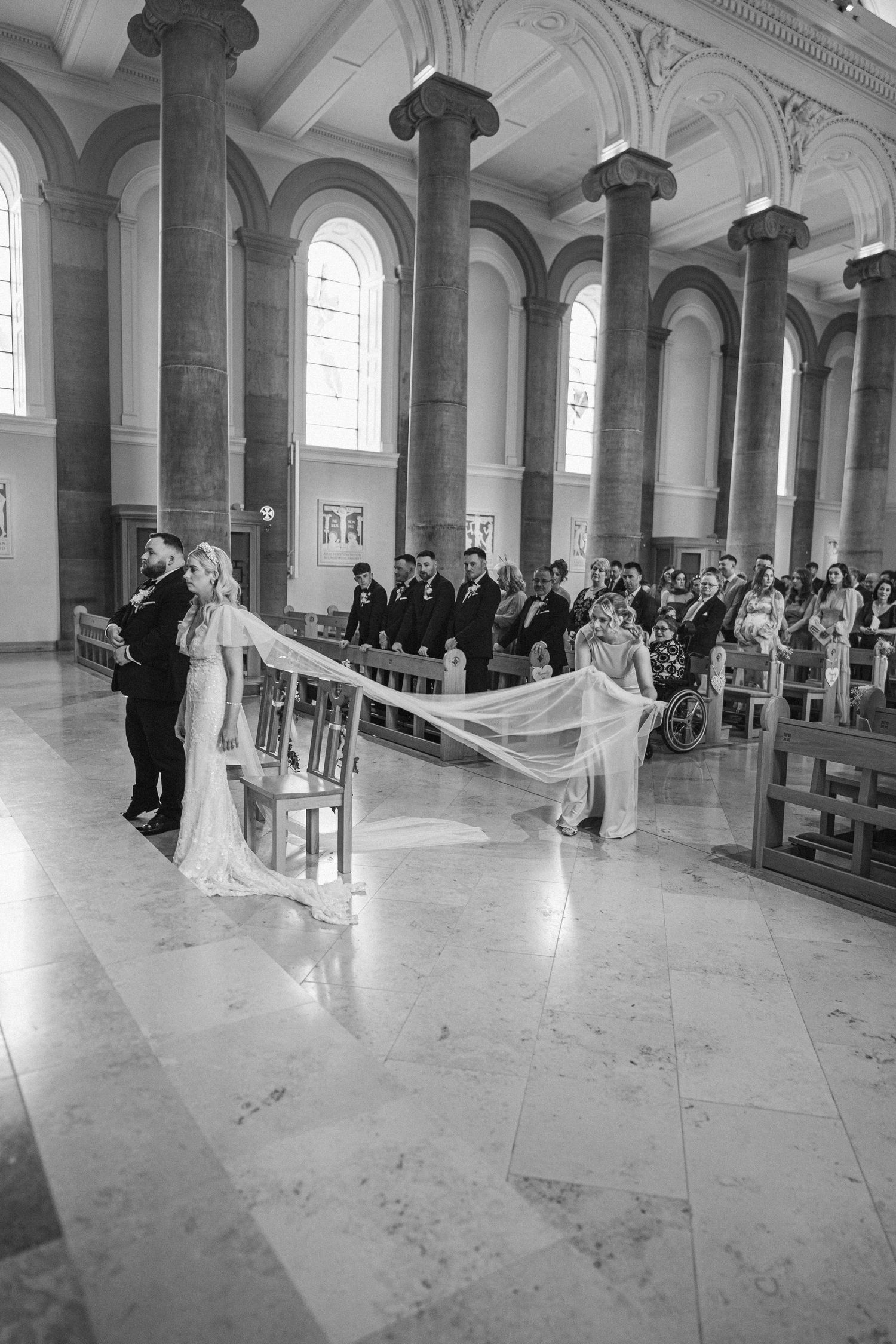 Black and white wide-angle photo of bride walking down cathedral aisle with long veil train at wedding ceremony.
