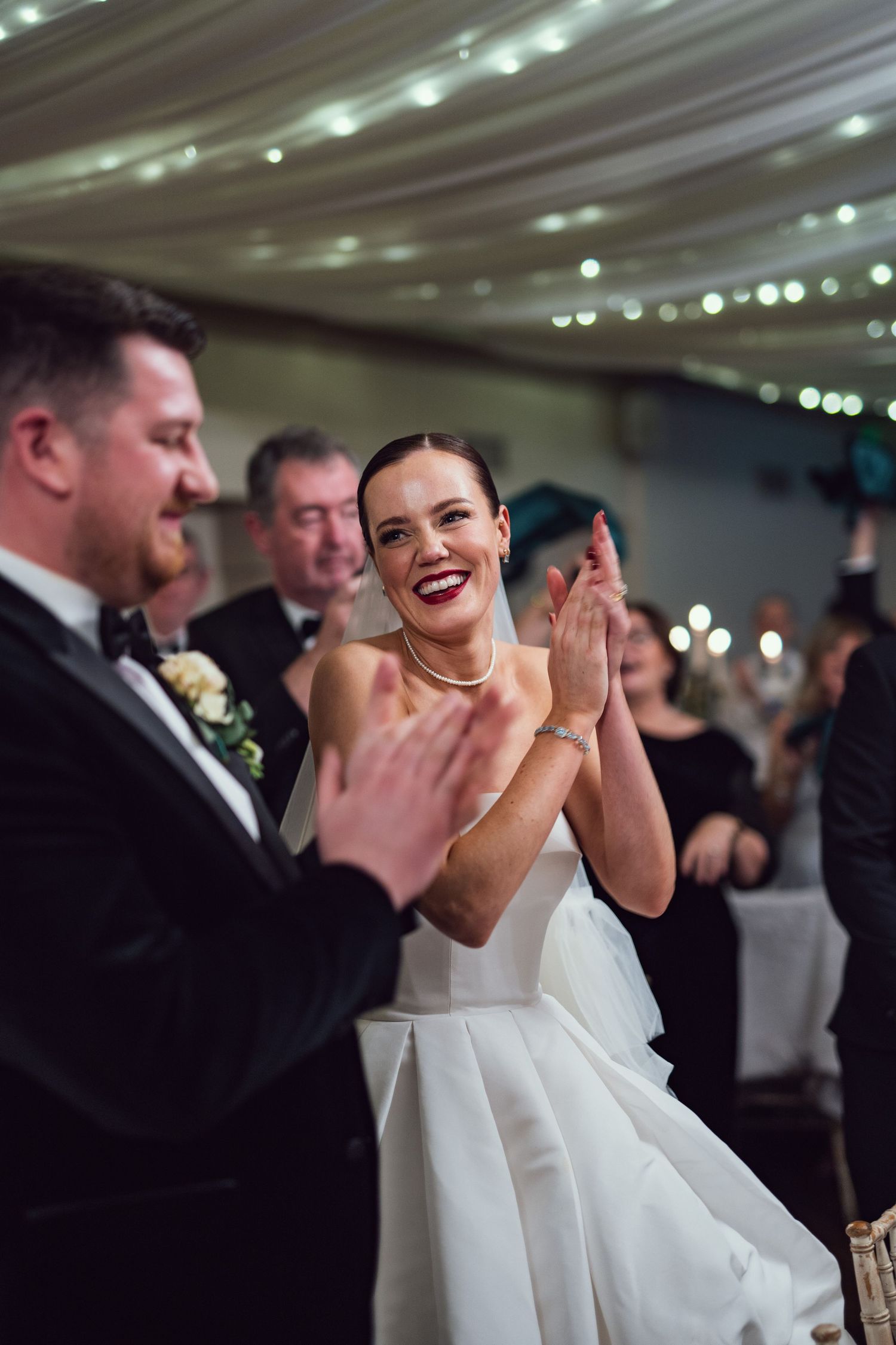 Radiant bride in strapless white gown clapping and smiling at wedding reception while seated beside groom in tuxedo.