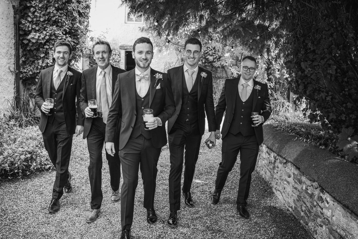 Black and white photo of a groom and four groomsmen walking confidently together holding drinks outdoors.