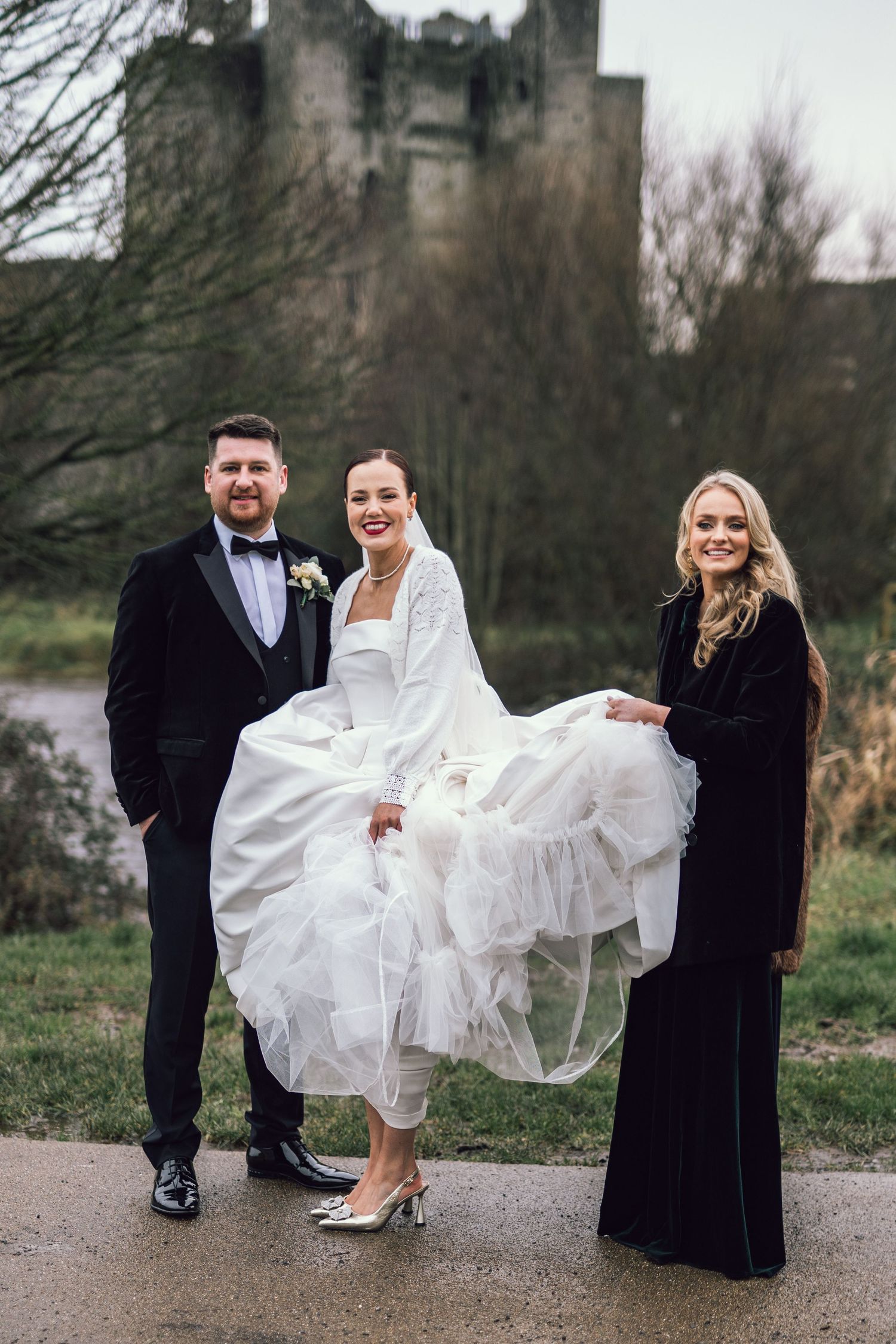 Bride in layered tulle gown poses with groom and bridesmaid in black dress before castle.