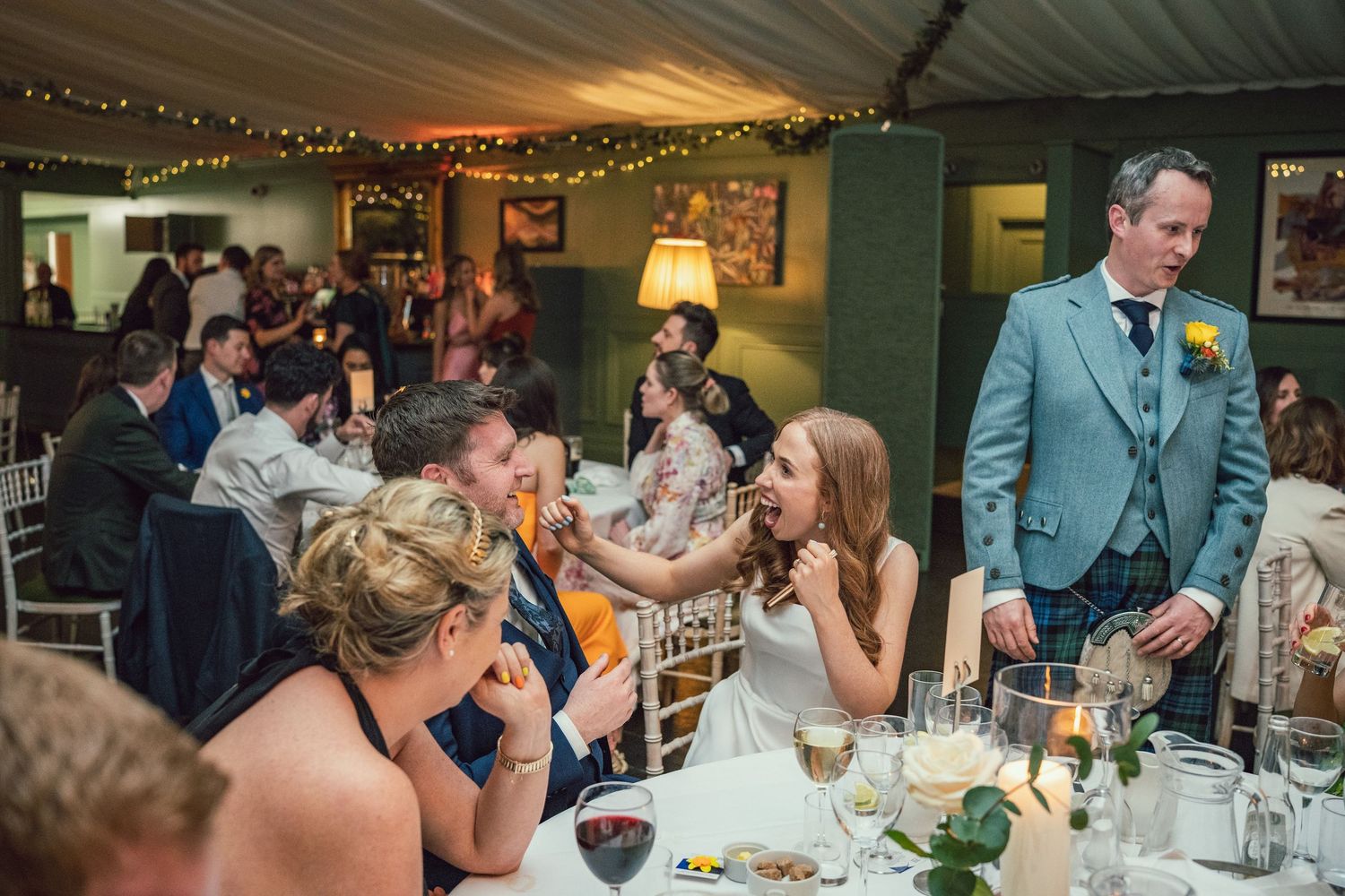 Wedding guests laughing and socializing at a candlelit reception table inside a decorated marquee venue.