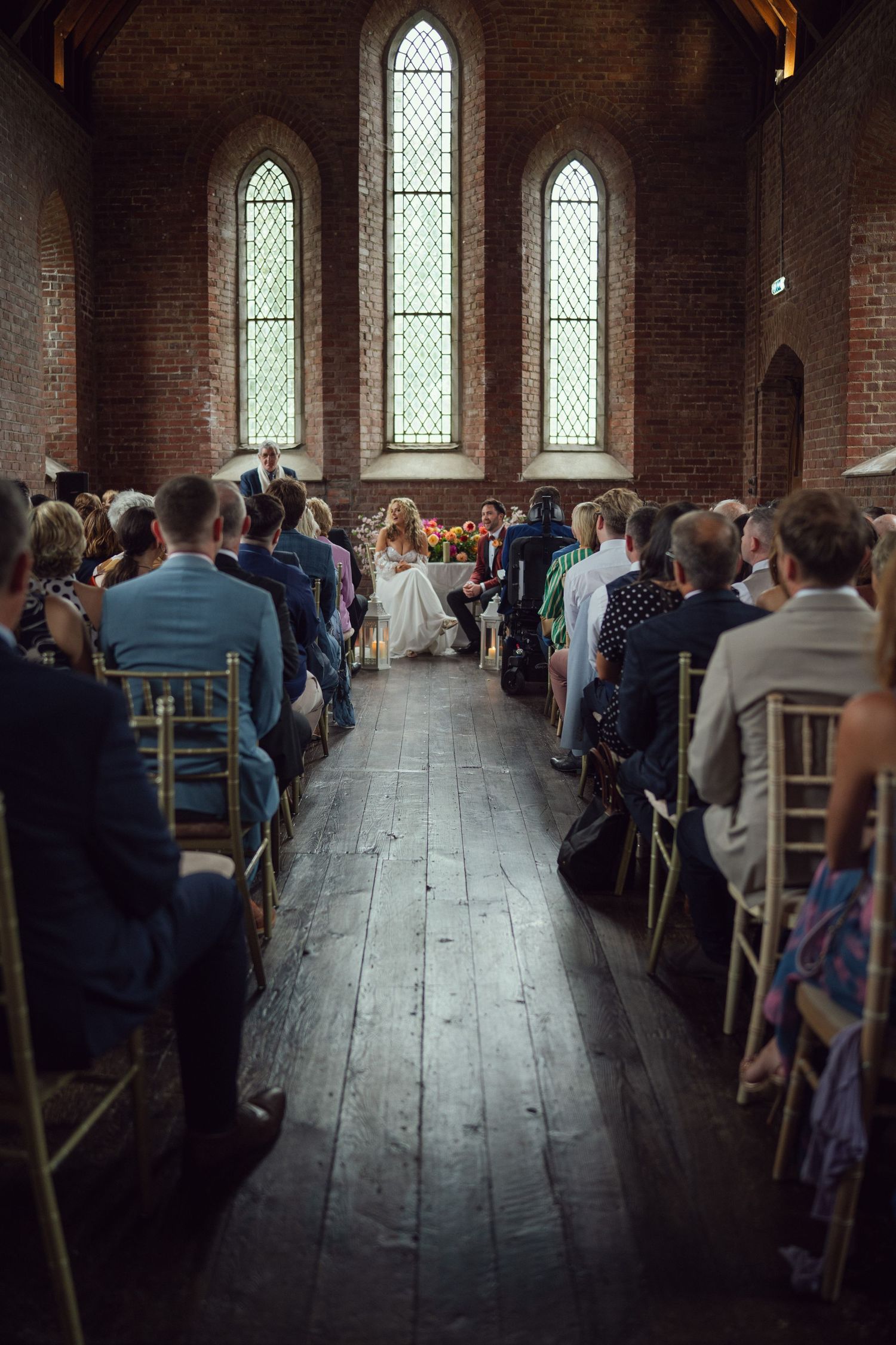 Wedding ceremony viewed from back at red chapel Kells in Co Meath with guests seated in gold chairs along wooden aisle.