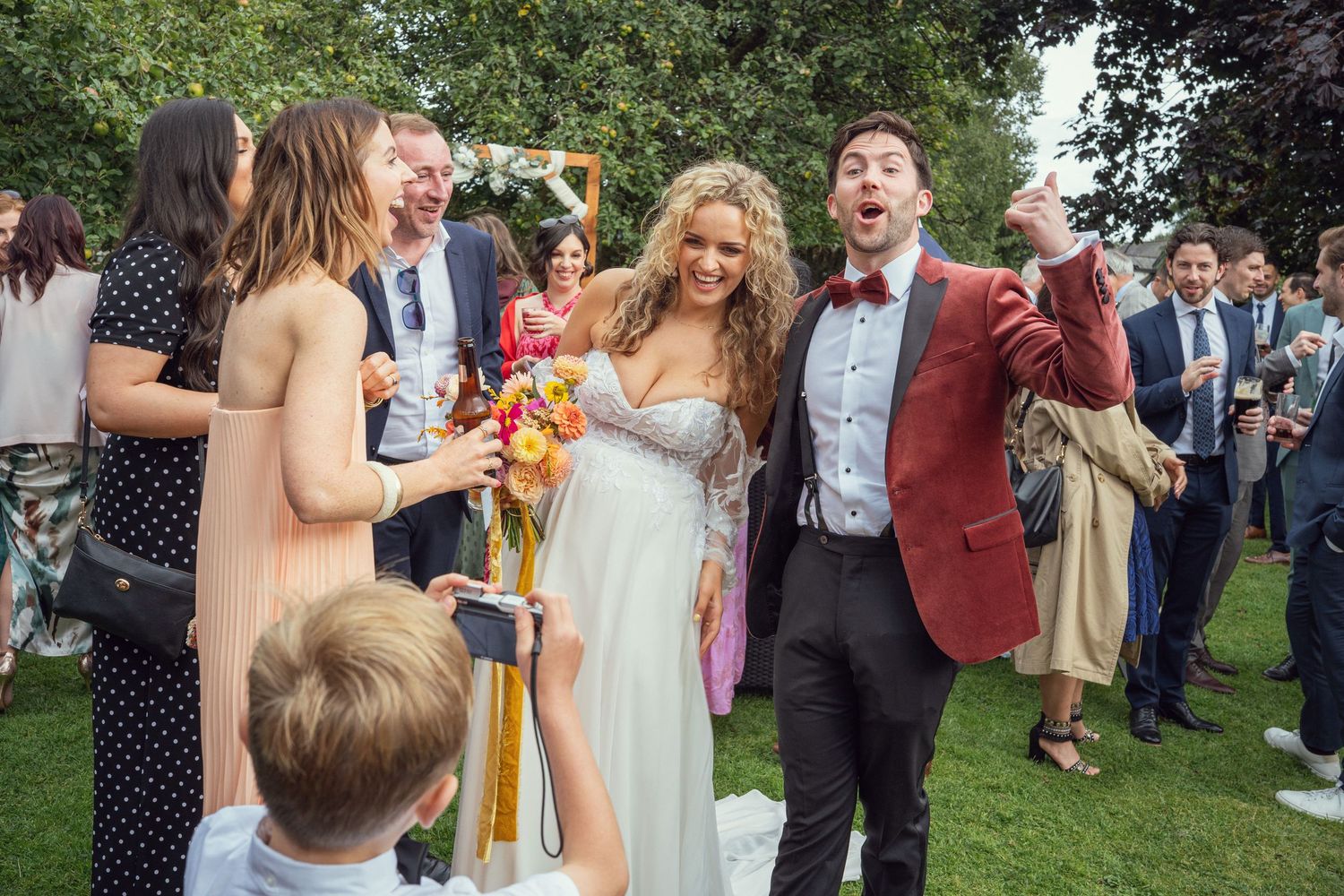 Bride in flowing gown and groom in red jacket celebrate joyfully with guests at outdoor garden wedding reception in Co Meath.
