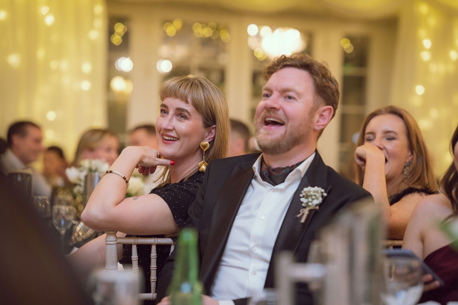 A couple laughing joyfully at a wedding reception dinner, surrounded by fairy lights and guests.