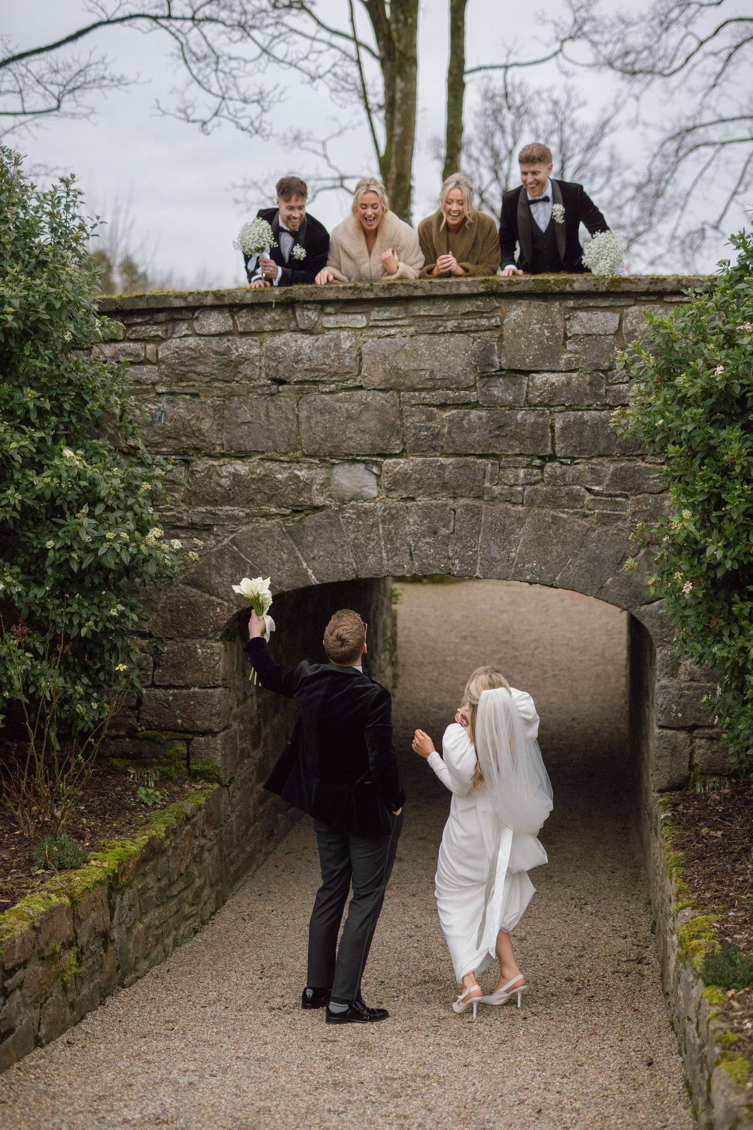Bride and groom walking through stone archway as wedding guests cheer from bridge above.
