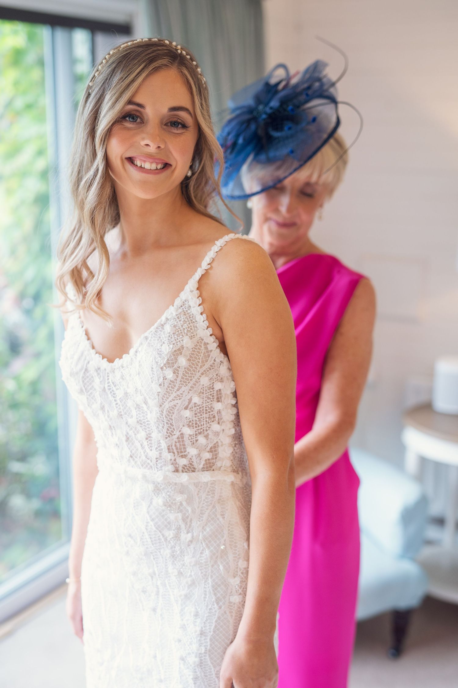 A smiling bride in a beaded gown stands as a woman in pink adjusts her dress before the wedding ceremony.