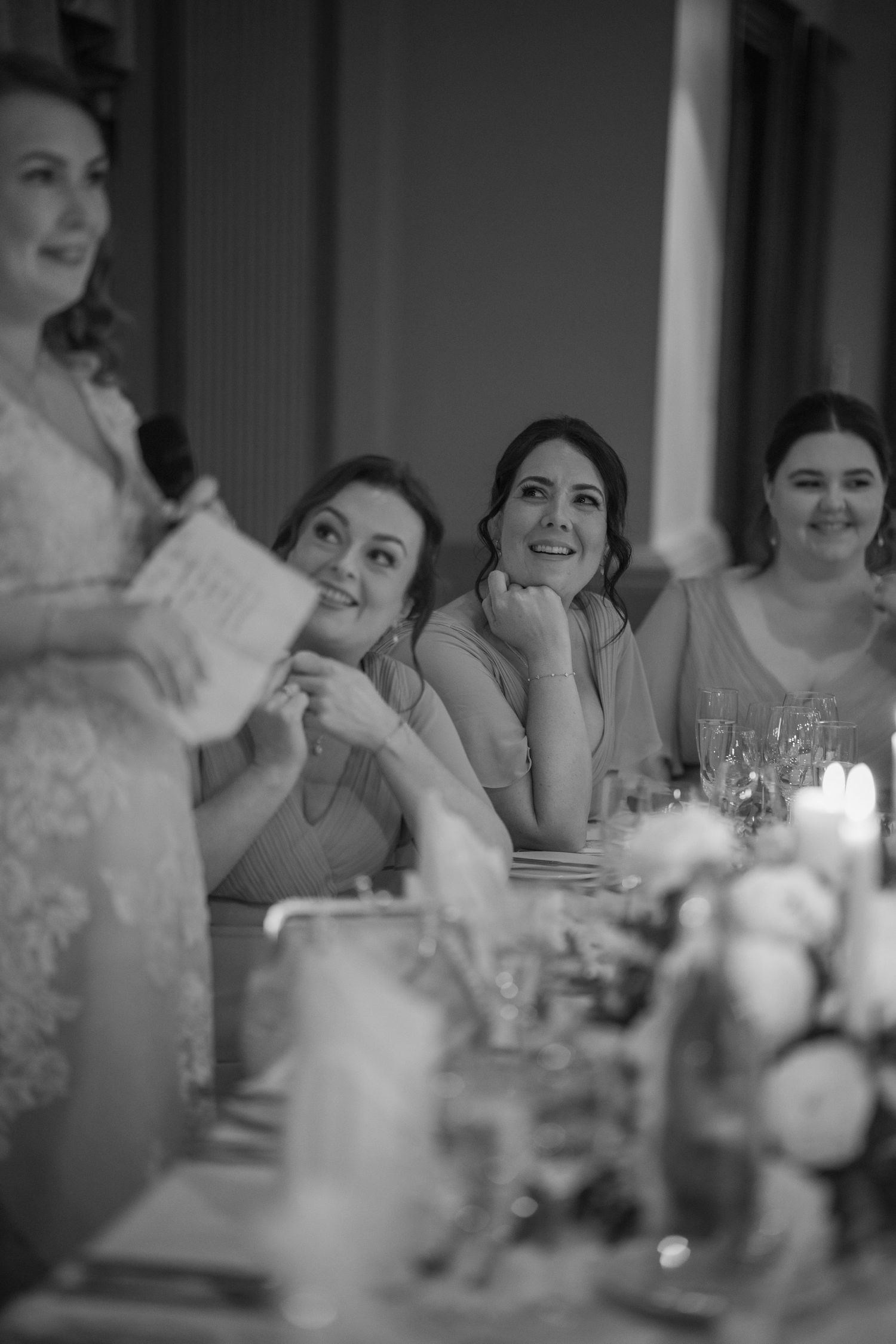 Black and white photo of bridesmaids smiling and laughing during wedding speech at reception table.