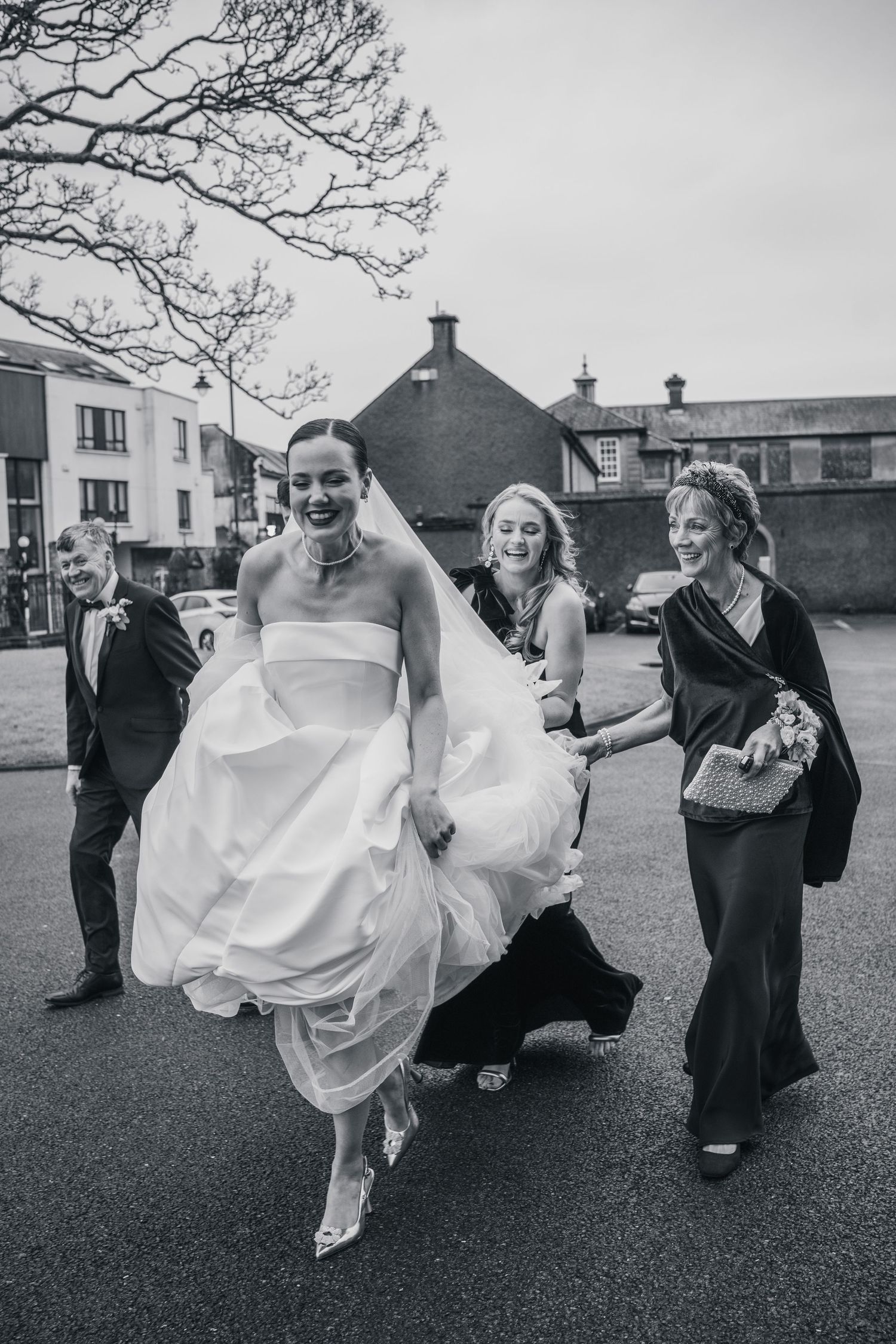 Laughing bride in strapless gown walks outdoors with bridesmaids lifting dress, black and white wedding photo.
