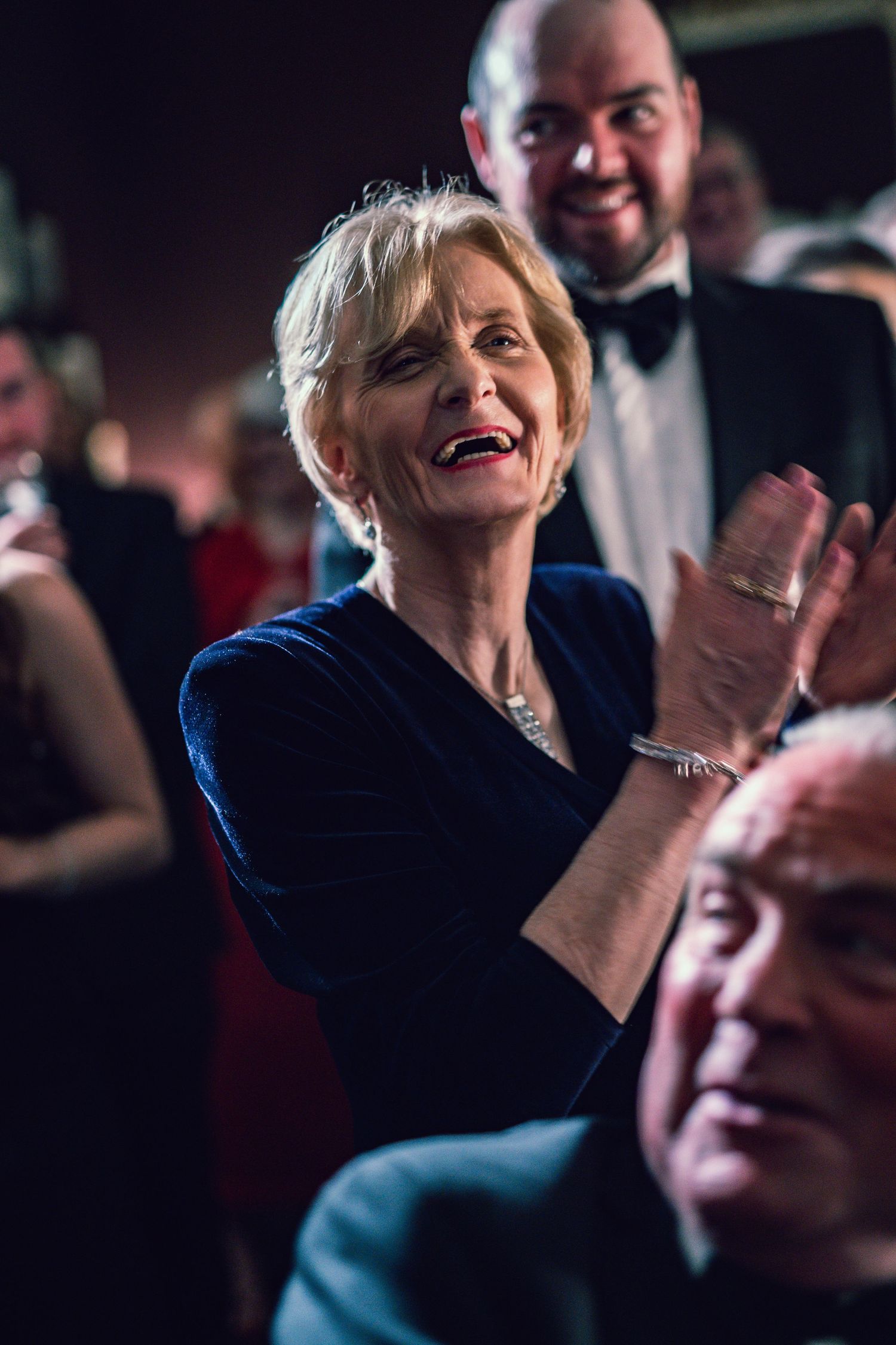 Elegantly dressed older woman in blue velvet applauds joyfully at a formal wedding reception event.