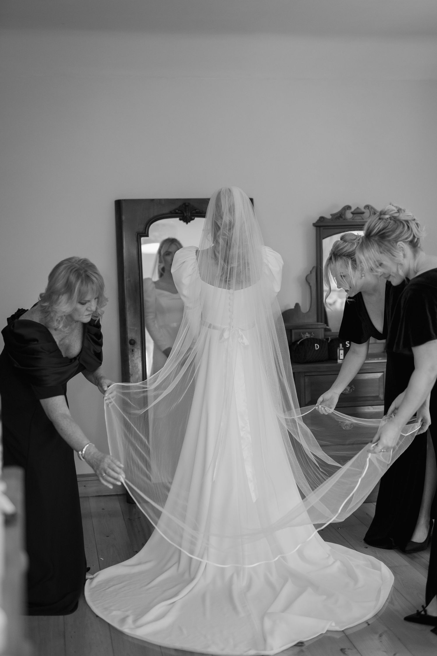 Two women in black dresses spread cathedral veil of bride from behind in black and white photo.
