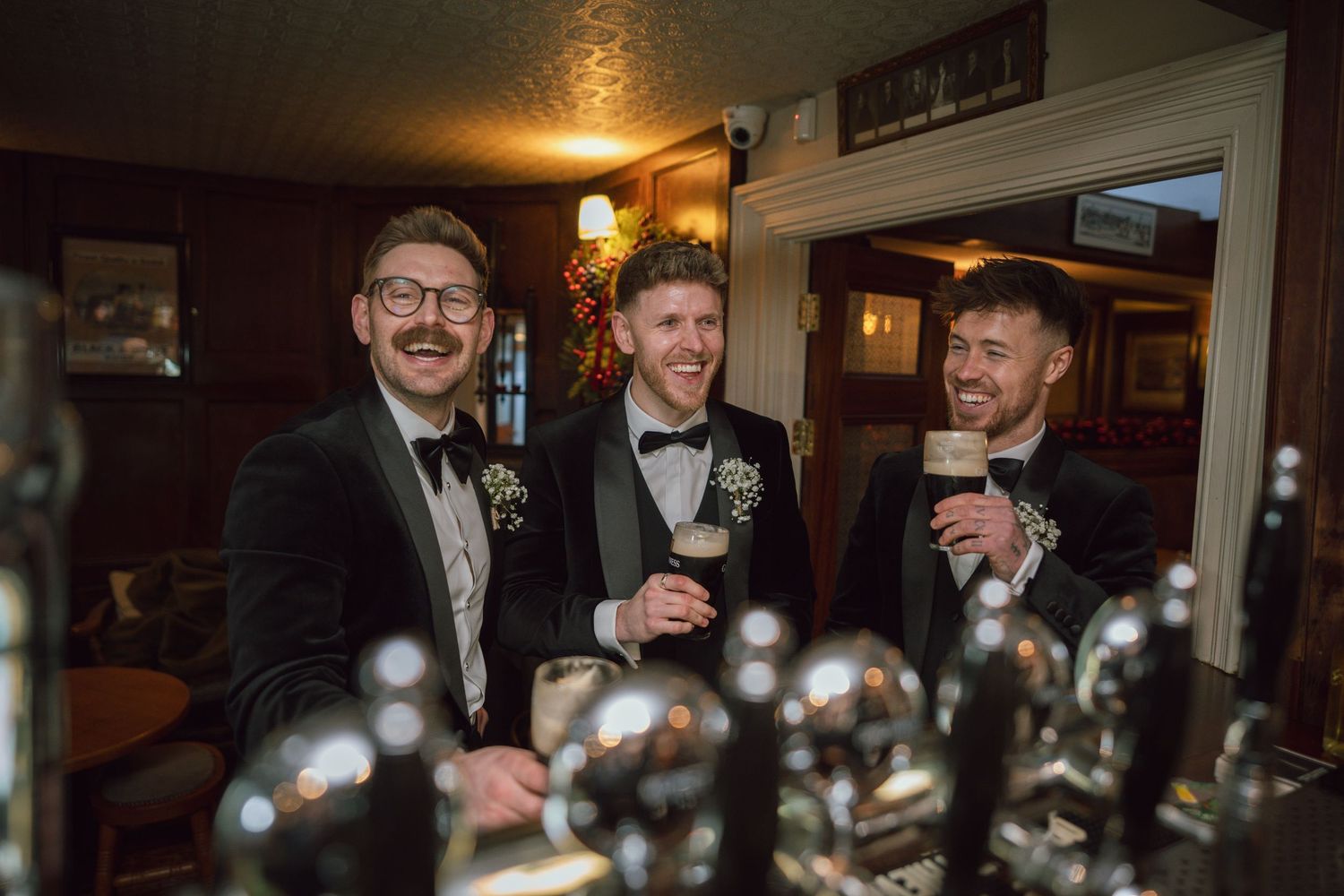 Three men in tuxedos smile and raise drinks at a pub bar during a wedding celebration.