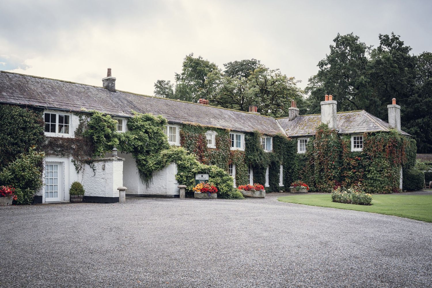Ivy-covered country house with colorful flower beds along the facade and a gravel driveway.