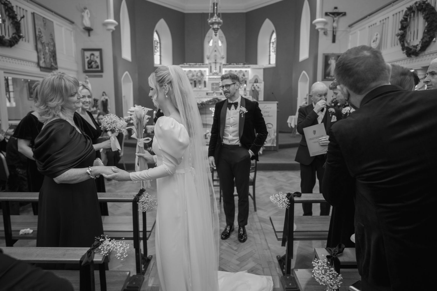 Bride and groom walk down church aisle together past seated wedding guests in black and white photo.