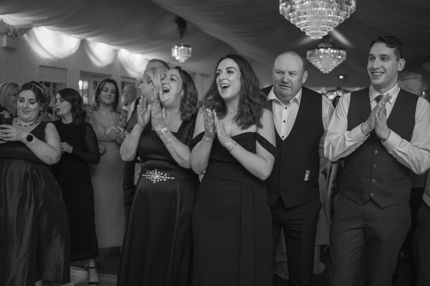 Wedding guests clap and celebrate on dance floor under chandelier at elegant reception.