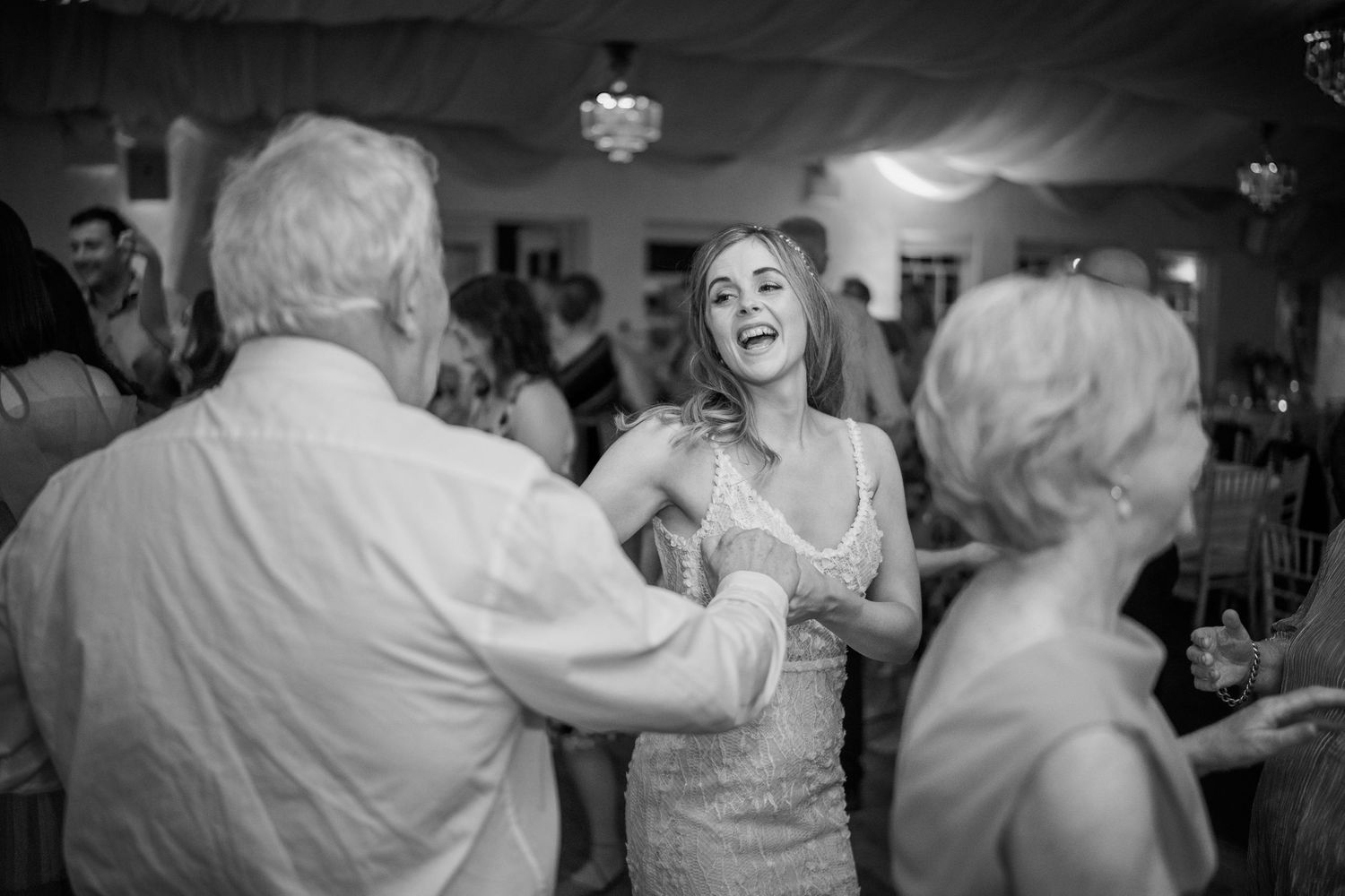 Black and white candid photo of a laughing bride dancing joyfully with older guests at her wedding reception.