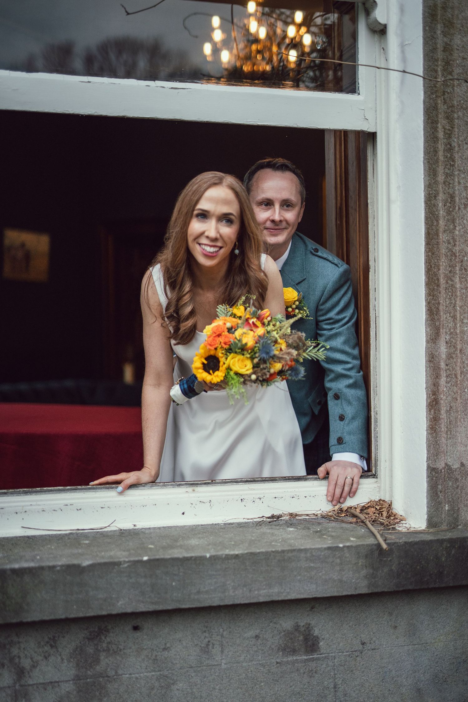 Bride and groom posing together in a window, bride holding a vibrant sunflower bouquet at their Scottish wedding.