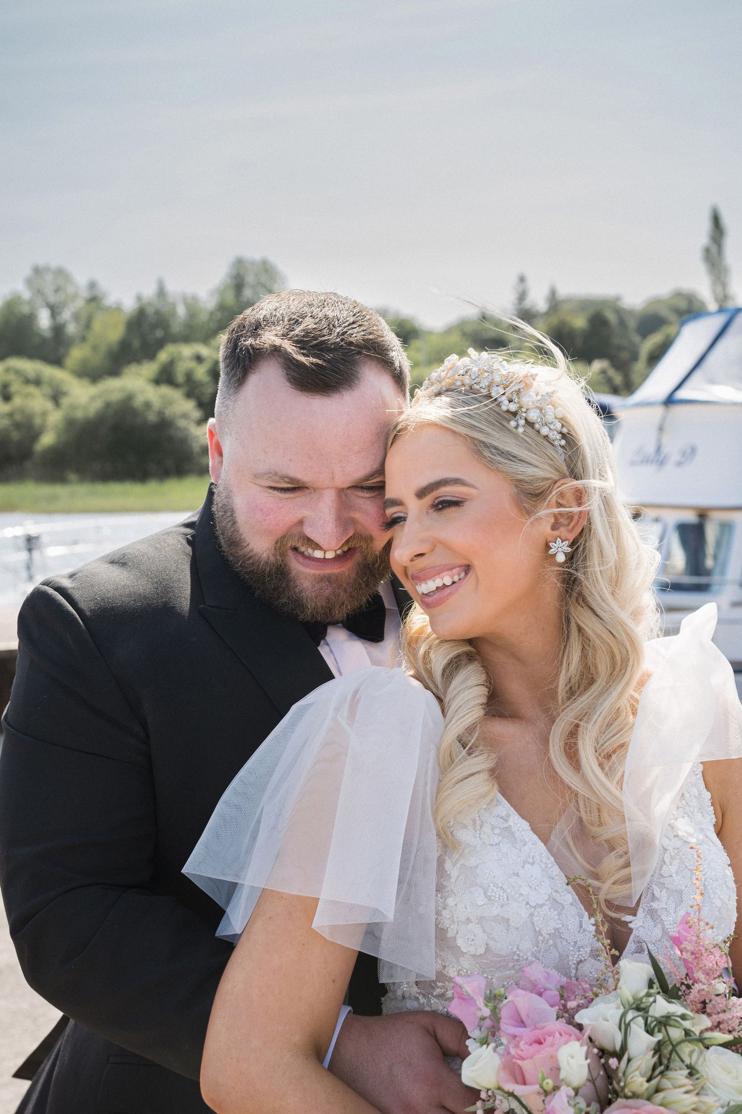 Groom nuzzling smiling blonde bride wearing floral headpiece outdoors near a marina on wedding day.