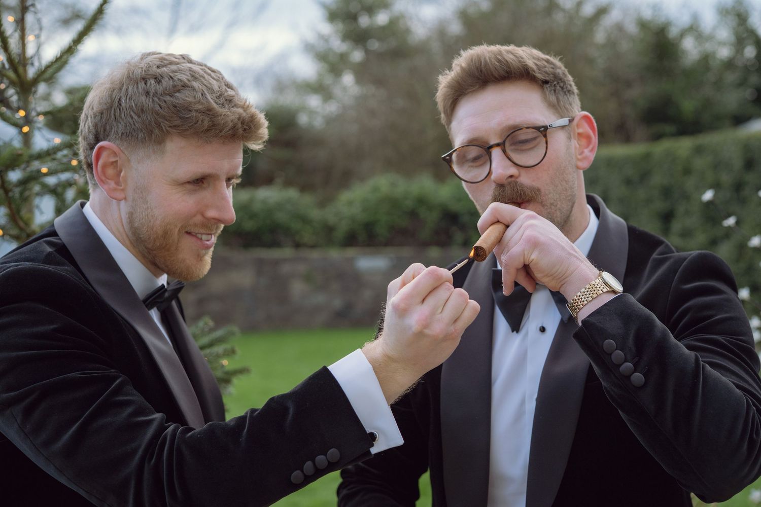 Two groomsmen in black tuxedos lighting a cigar together outdoors at a wedding.