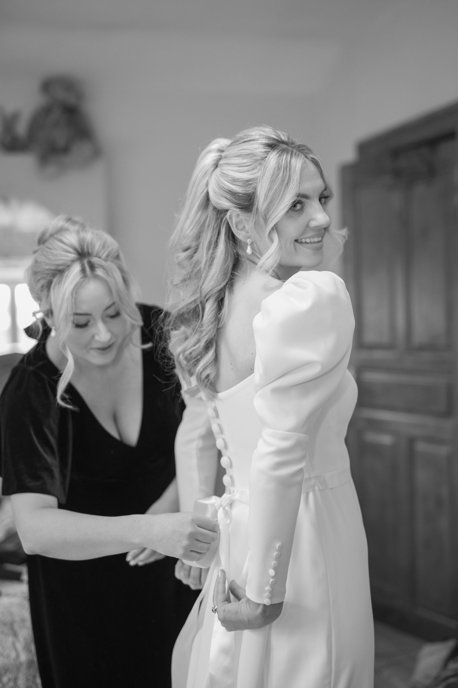 Bridesmaid in black velvet helps smiling bride with buttons on white wedding dress back.
