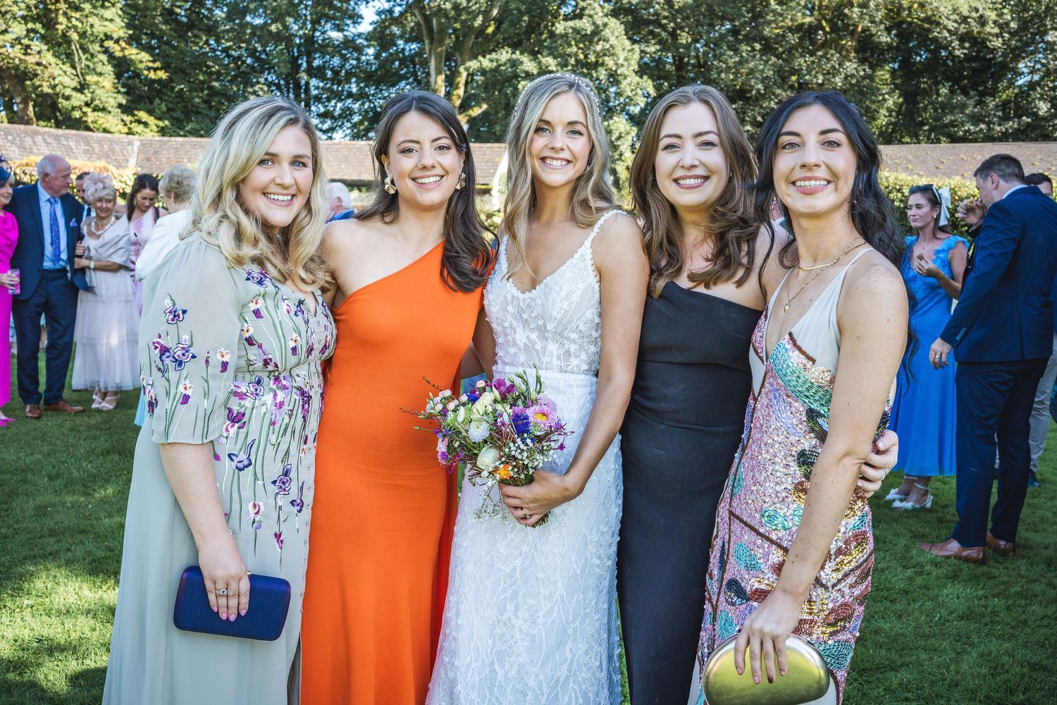 Bride in lace gown poses with four female wedding guests on sunny lawn.