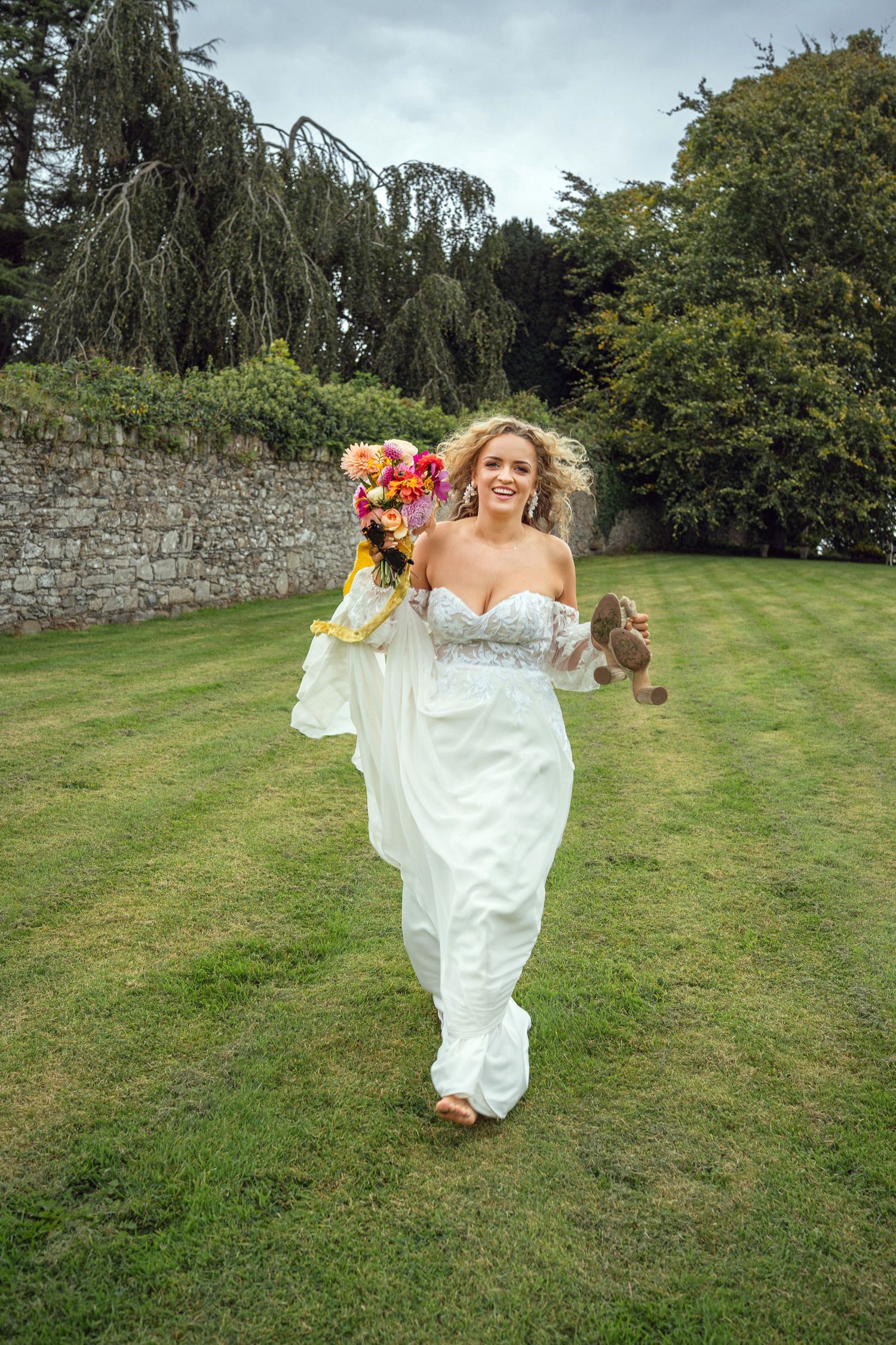 Joyful bride in white lace off-shoulder gown running on lawn holding colorful bouquet and shoes outdoor garden wedding reception in Co Meath