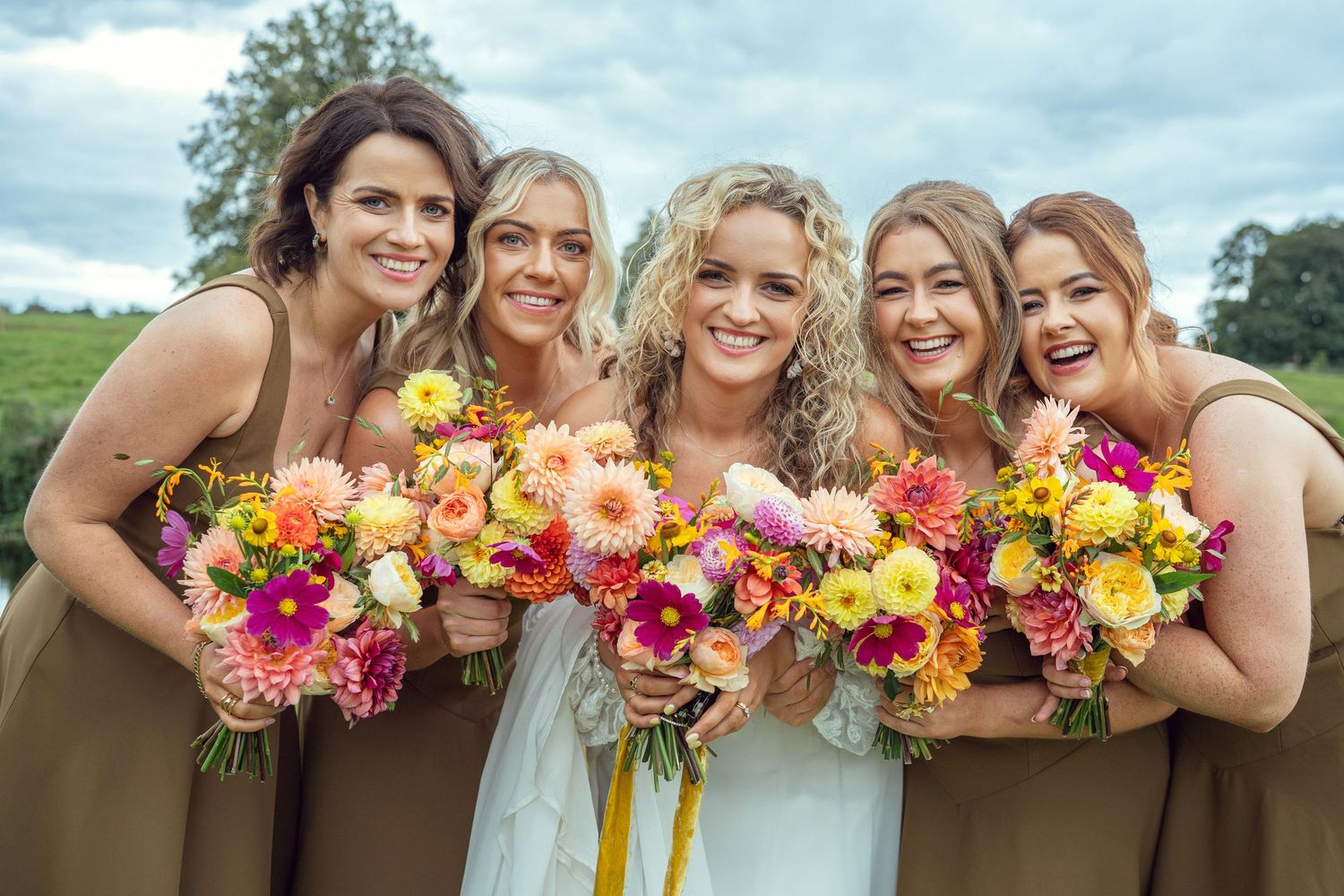Bride with four bridesmaids in tan dresses holding vibrant flowers outdoor garden wedding reception in Co Meath.