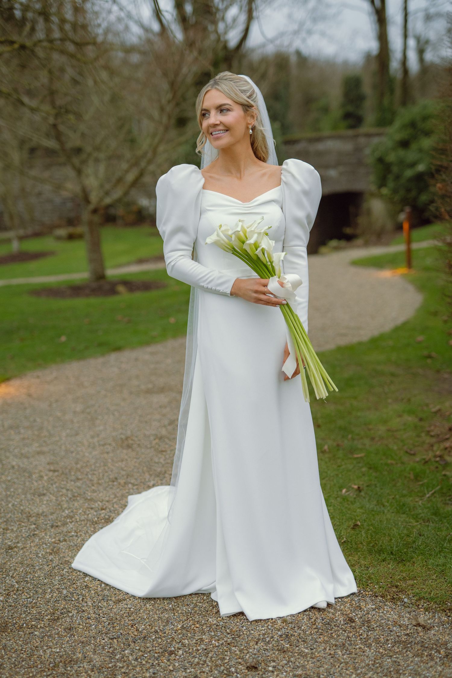 Elegant bride in long-sleeved white gown with puff shoulders holds calla lily bouquet in garden setting.