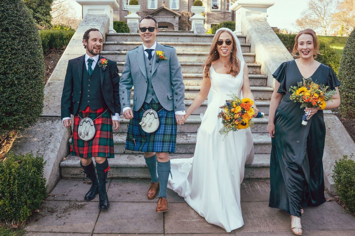 Wedding party with kilted groomsmen, bride holding sunflowers, and bridesmaid on steps.