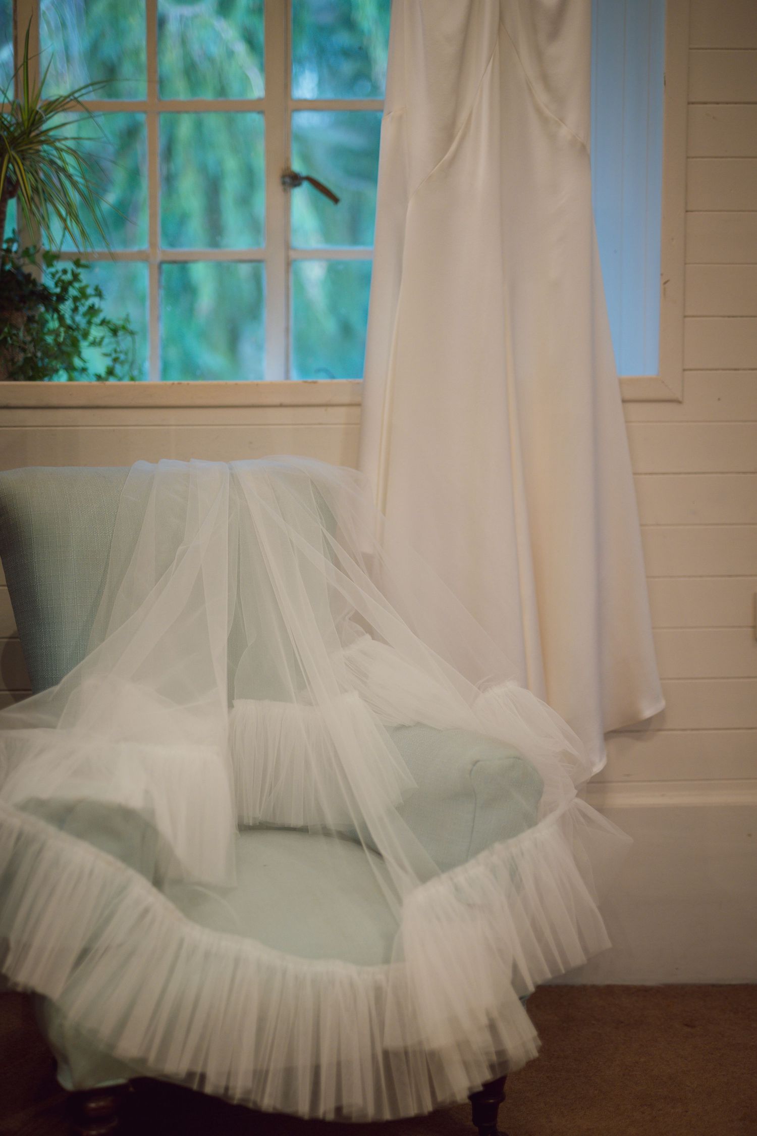 Wedding veil draped over vintage chair near window with bridal gown.