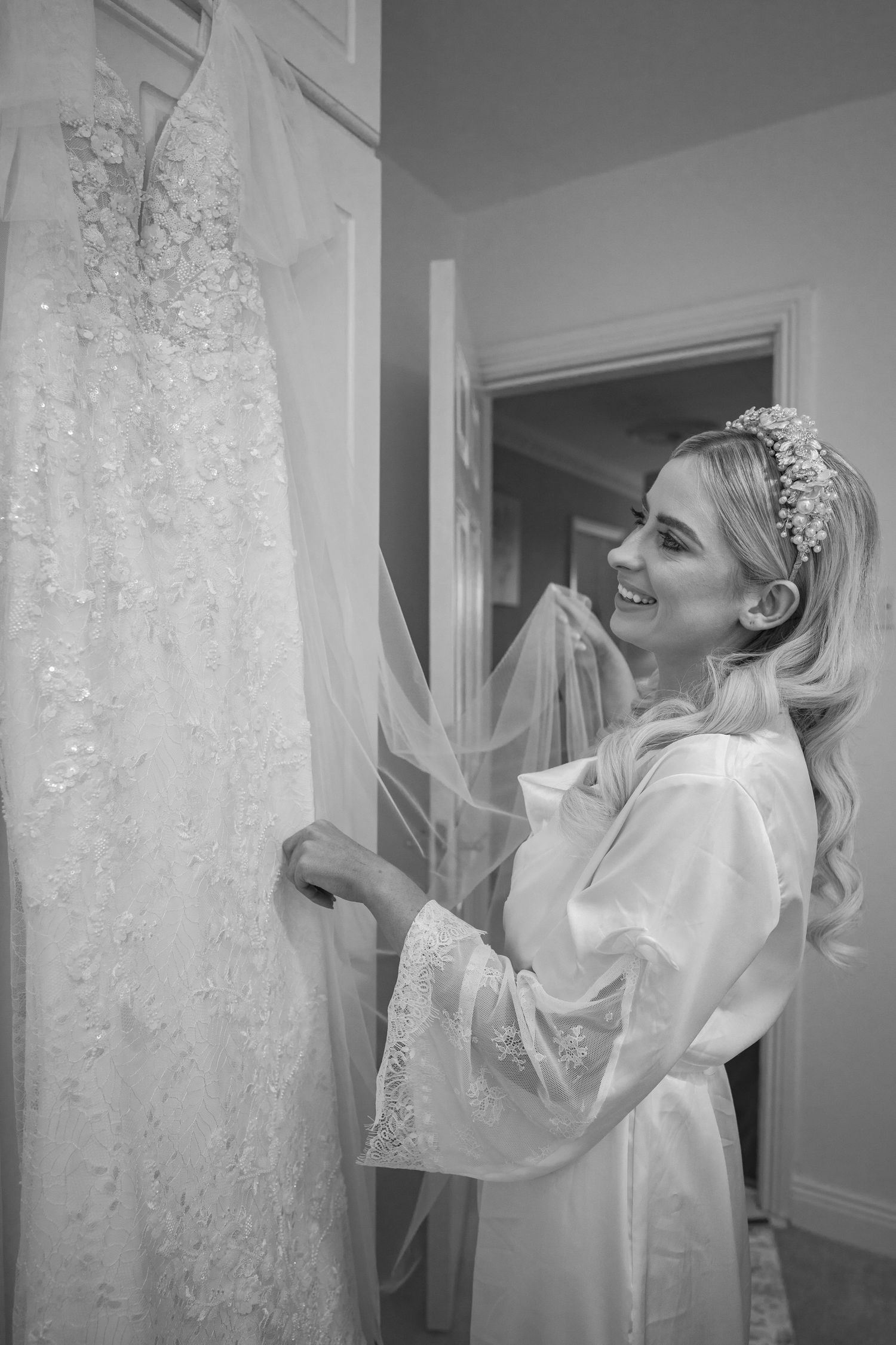 Black and white photo of smiling bride in robe admiring her lace wedding dress and veil.