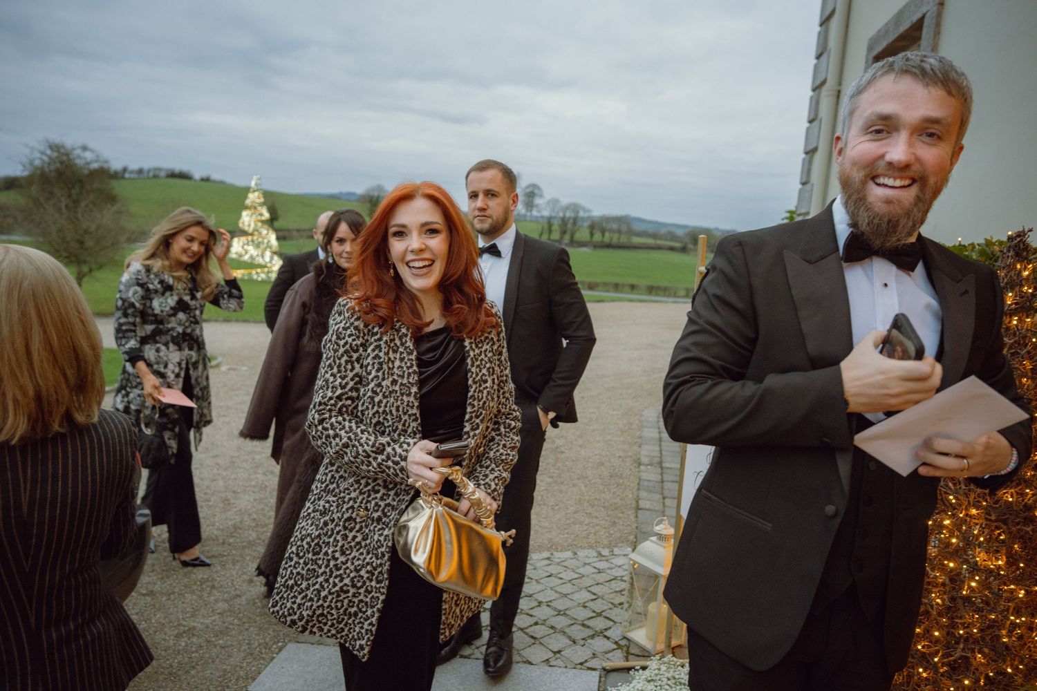 Guests arrive at a wedding venue, smiling and dressed in formal attire outdoors.