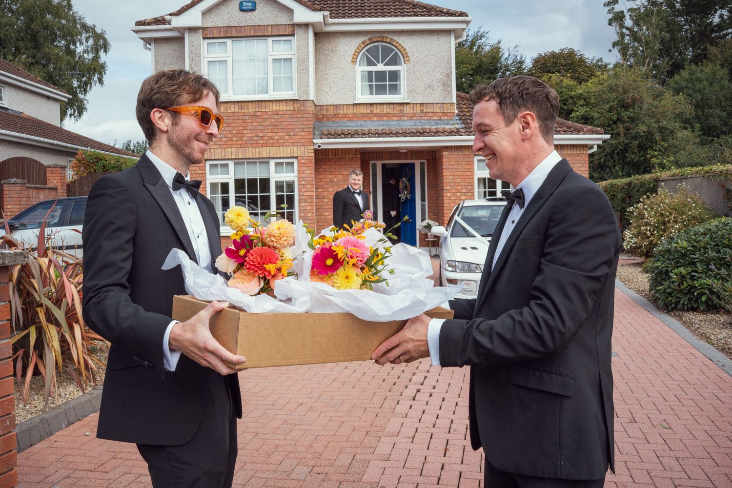 Two men in black tuxedos exchange colorful wedding bouquets outside a suburban house on wedding day.