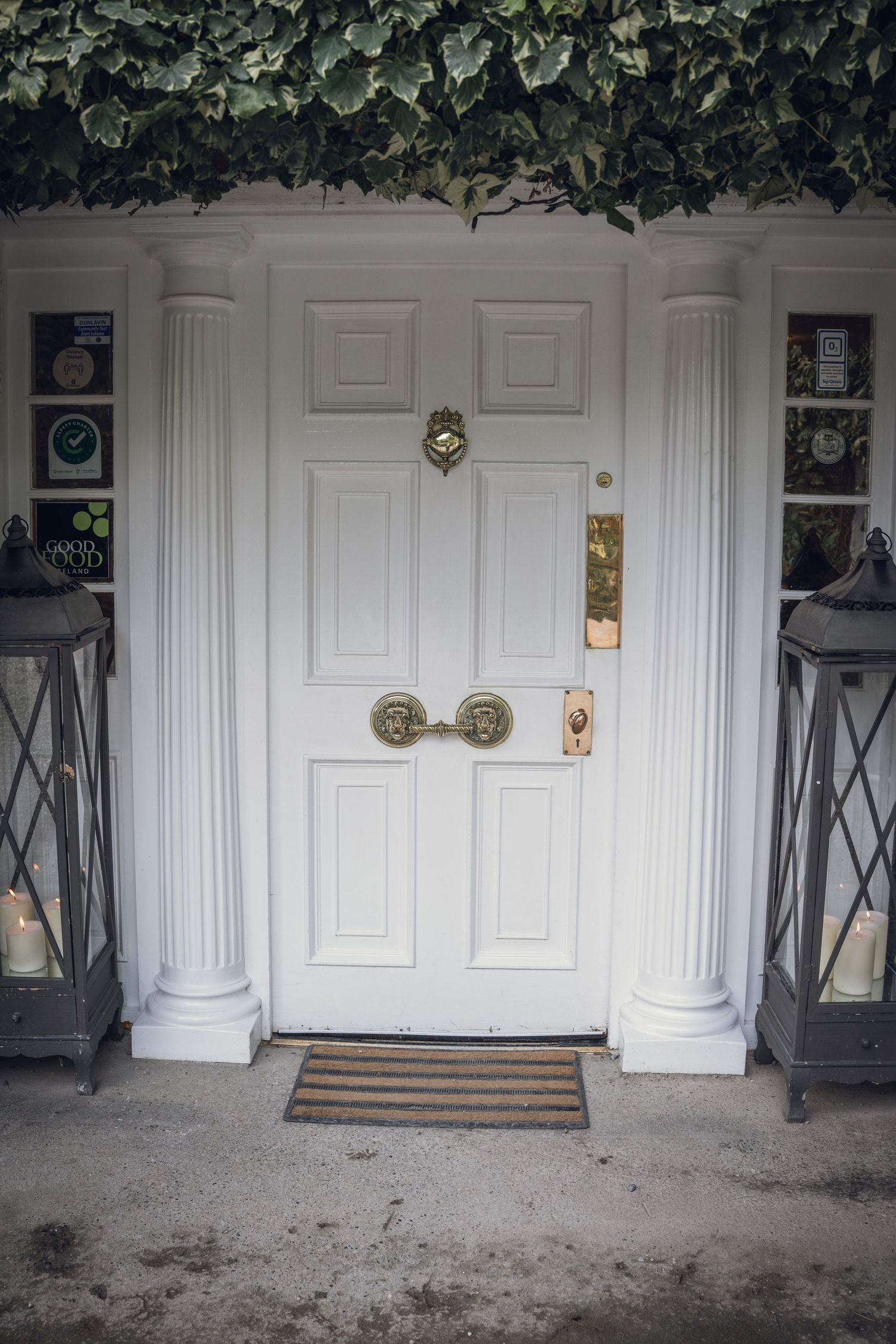 Elegant white front door with brass knocker flanked by pillar columns and lanterns with ivy overhead.