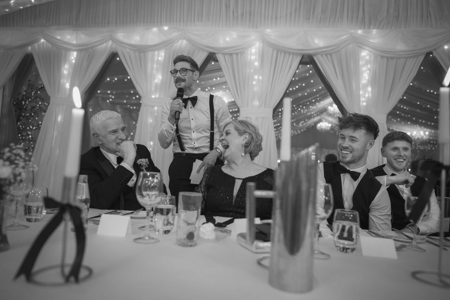A man gives a speech at a wedding reception in a fairy-lit marquee while guests laugh and celebrate.