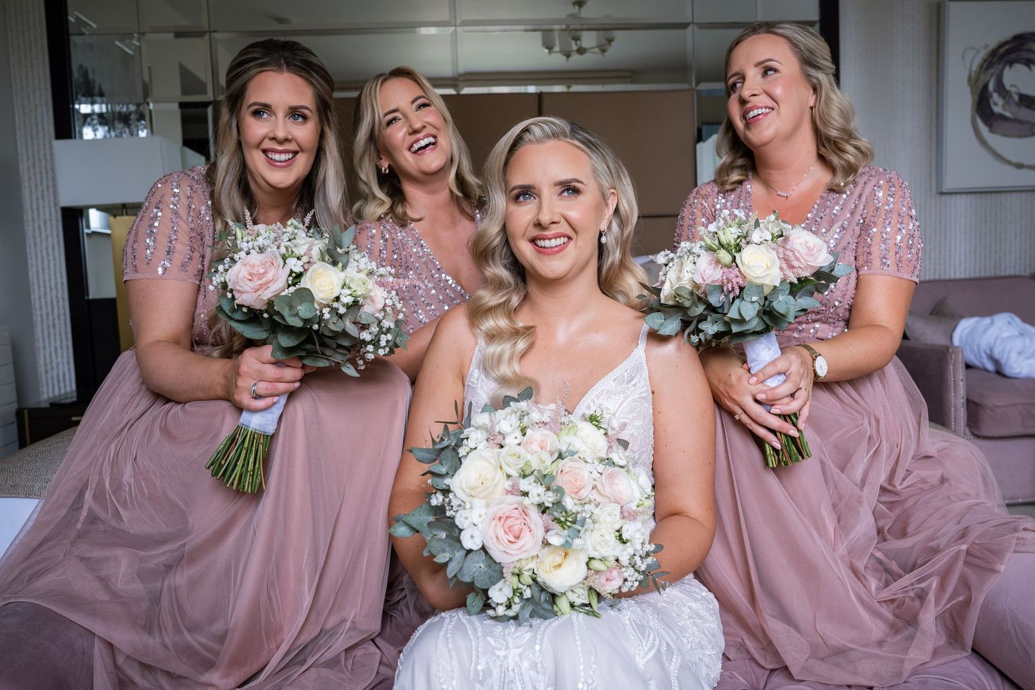 Radiant bride in white gown poses with three bridesmaids in pink sequin dresses holding bouquets.
