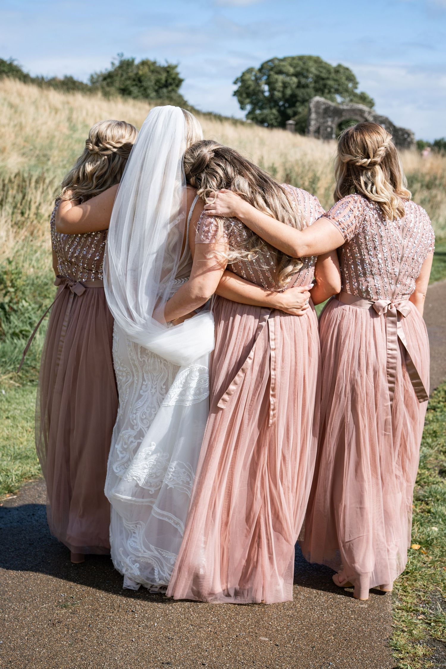 Bride in white lace gown and three bridesmaids in dusty rose dresses embrace from behind outdoors.