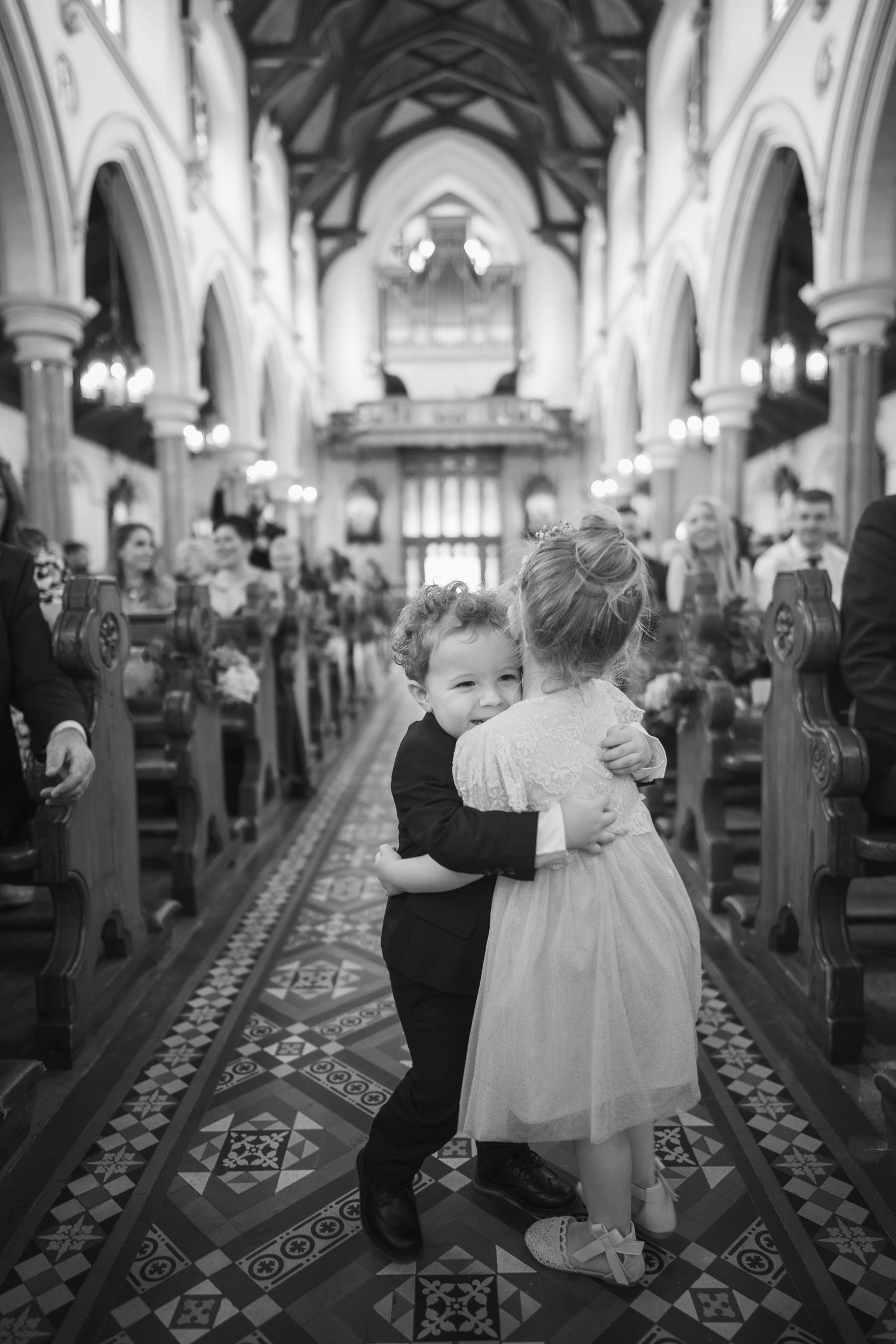 Two young children in wedding attire hugging each other in the aisle of a grand church.