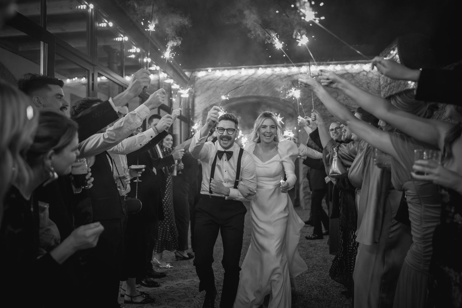 Bride and groom walking through sparkler tunnel surrounded by cheering wedding guests at night.