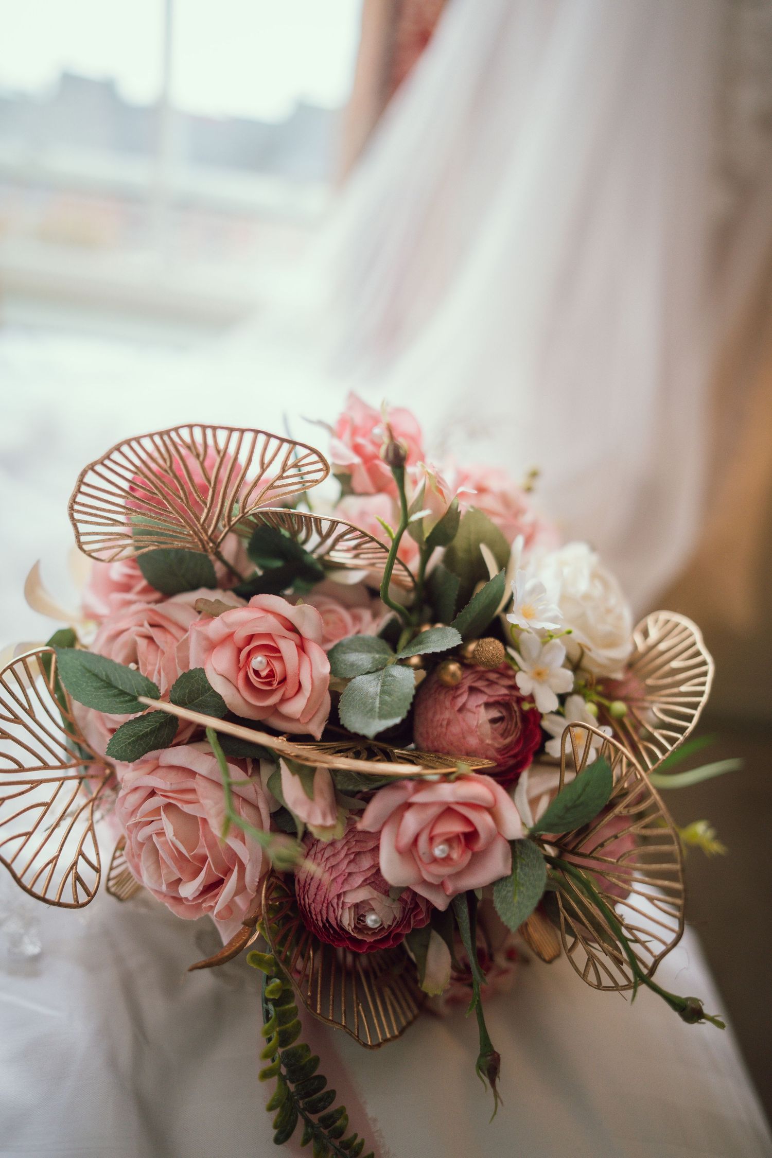 Bride holding romantic pink rose bridal bouquet with gold butterfly accents near a sunlit window.