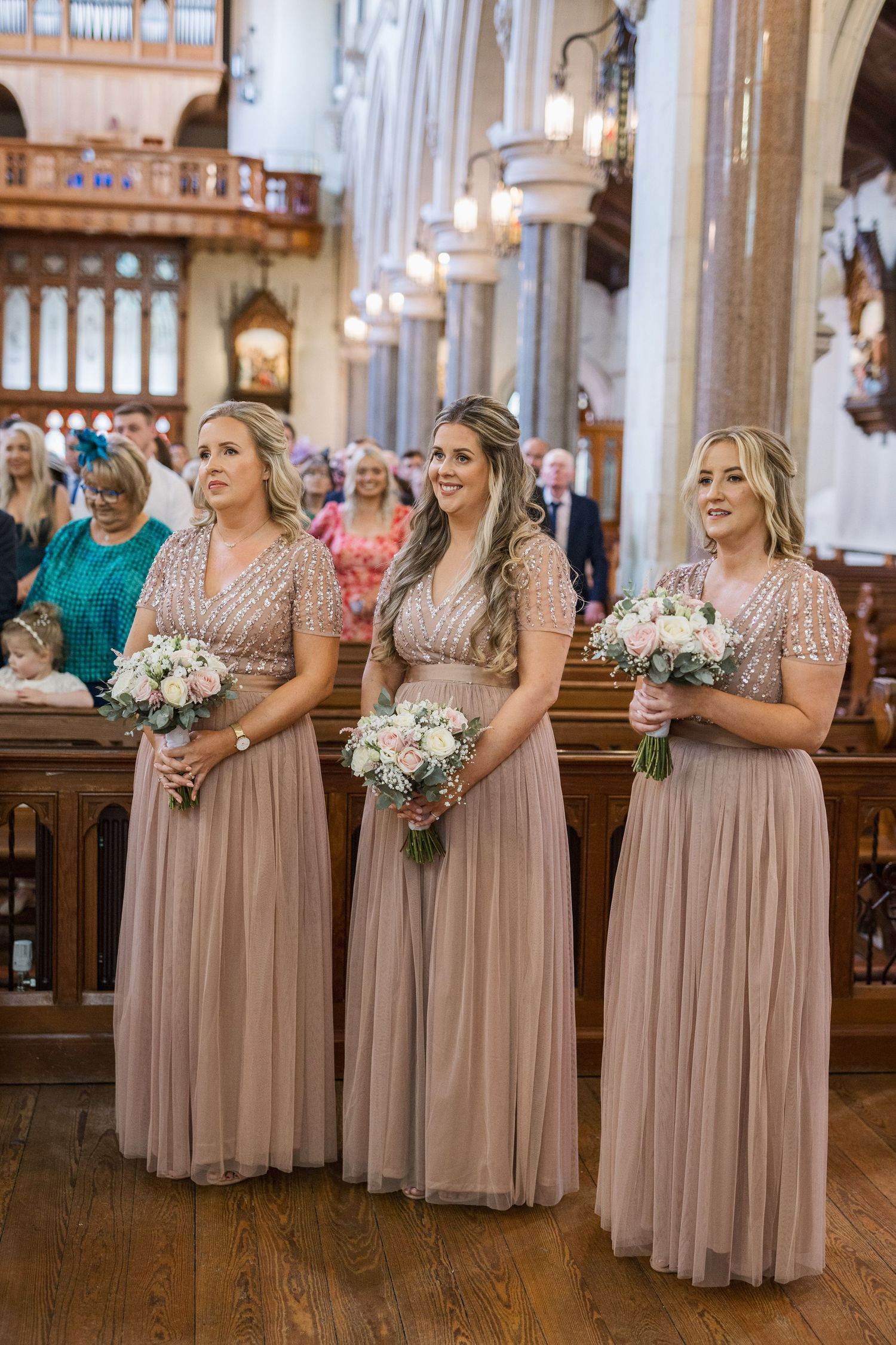 Three bridesmaids in dusty rose sequin and tulle gowns hold white floral bouquets inside a church.