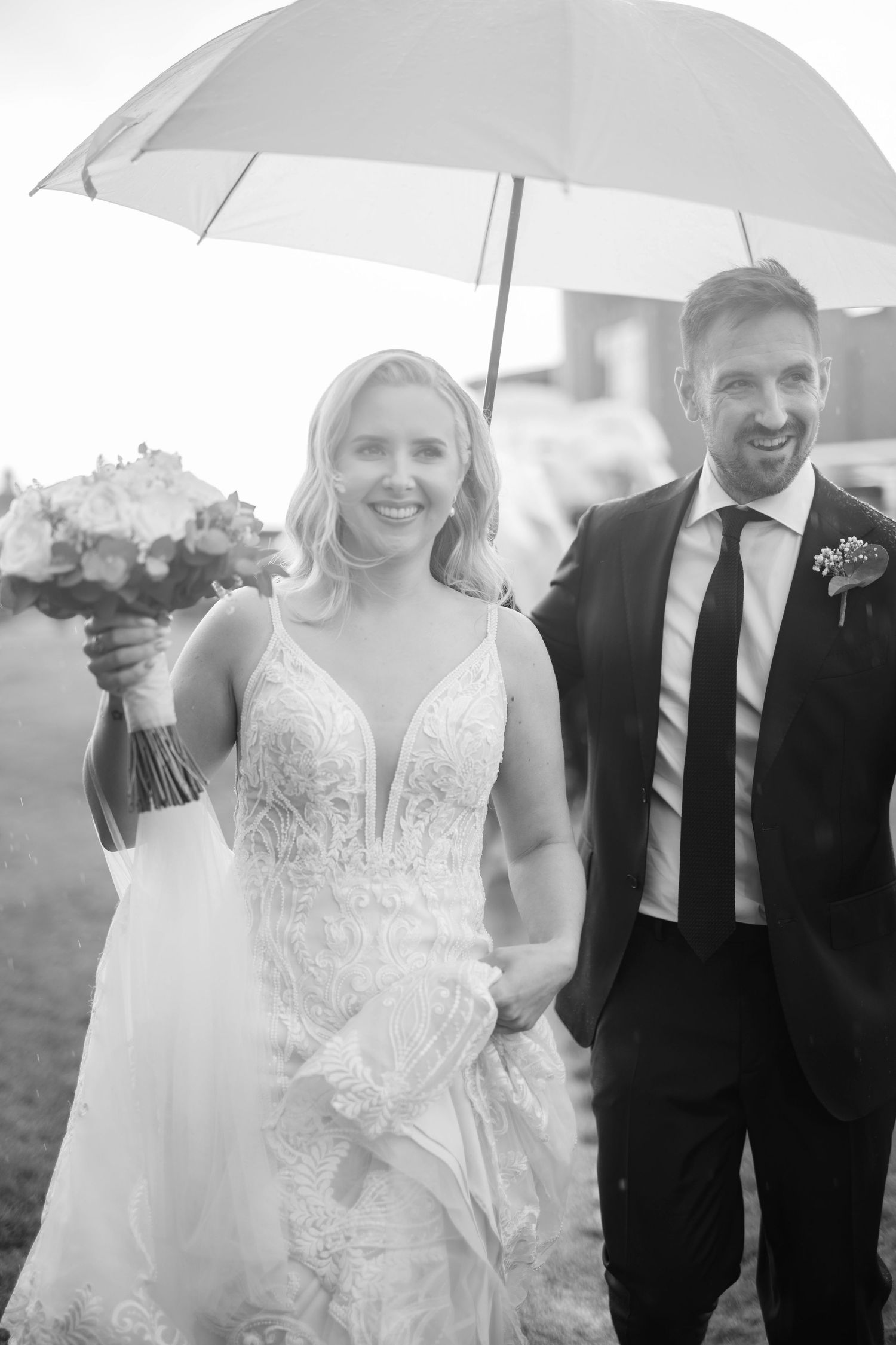 Black and white photo of smiling bride and groom walking together under an umbrella in the rain.
