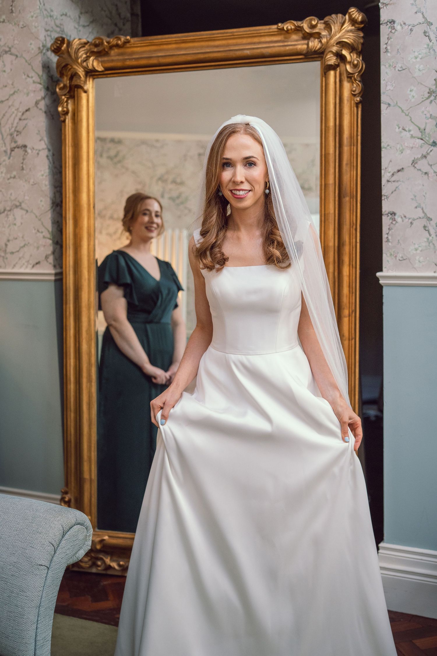 Elegant bride in minimalist white satin gown and veil posing before a ornate gold mirror with bridesmaid behind her.
