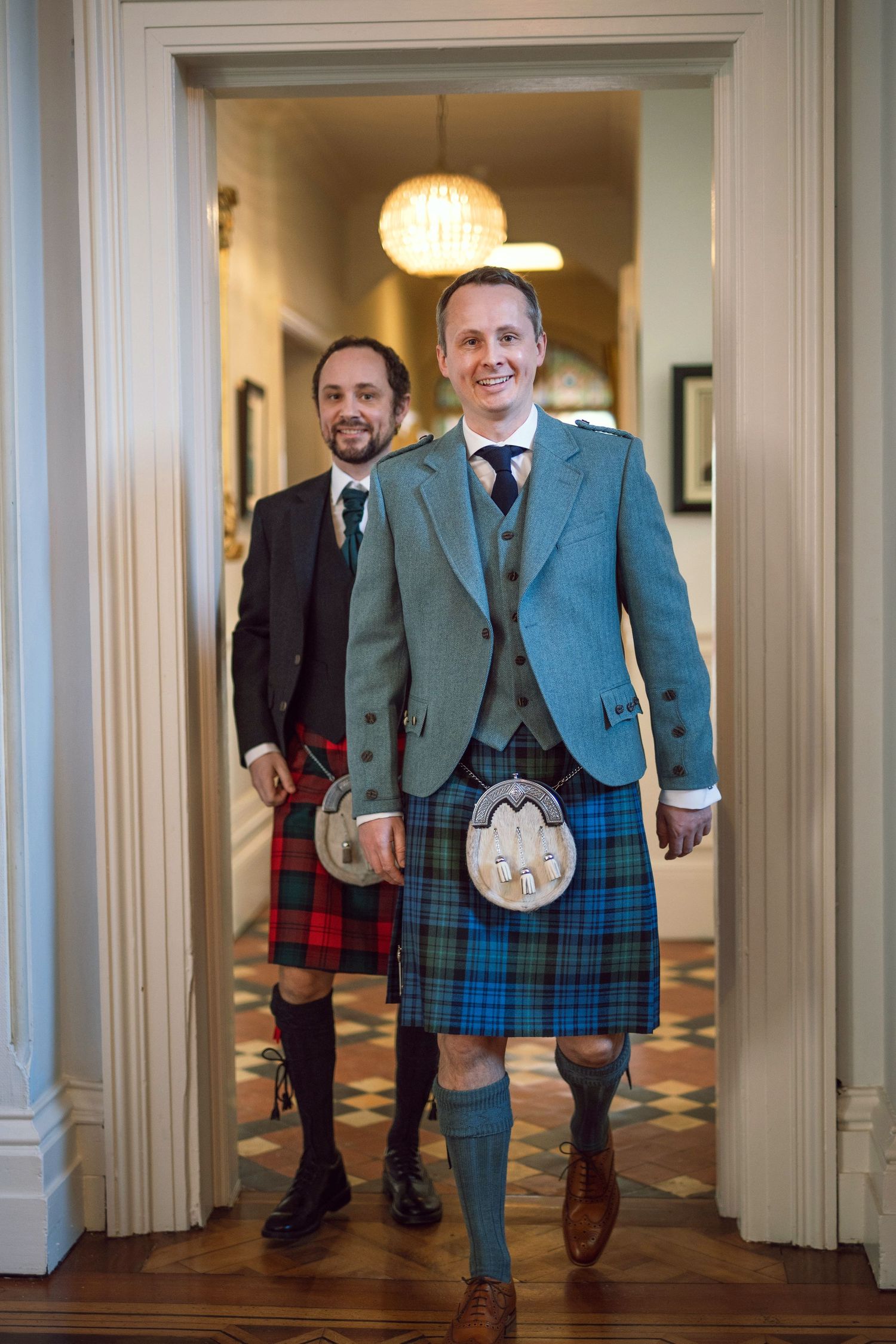 Two men in traditional Scottish kilts smiling while walking through a doorway.
