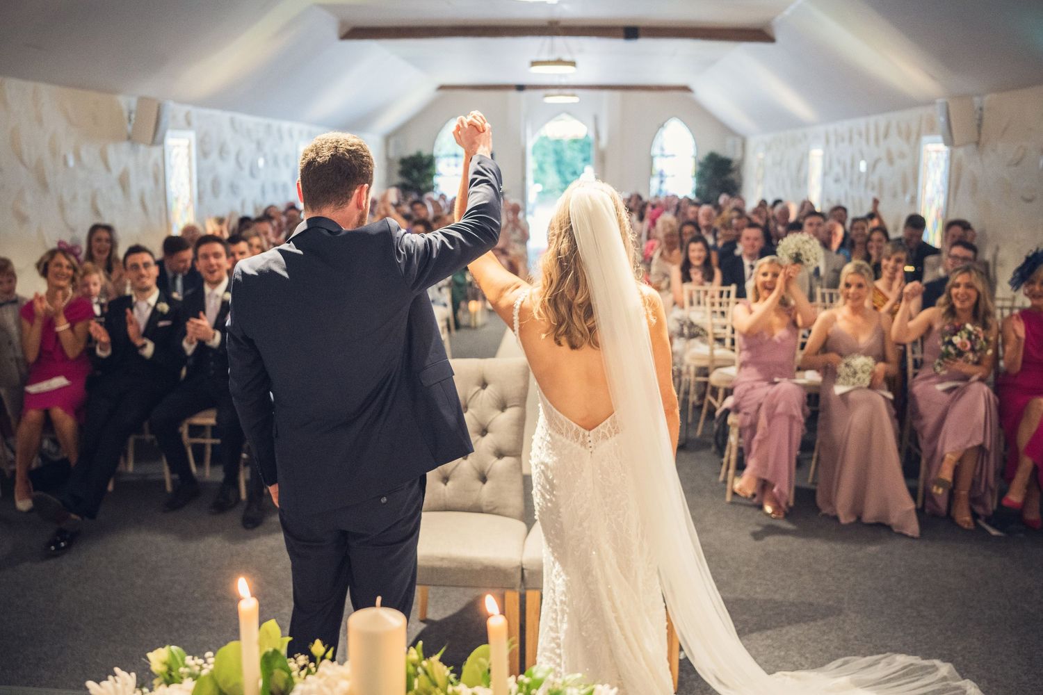 Bride and groom celebrate at altar as guests cheer in elegant white chapel.